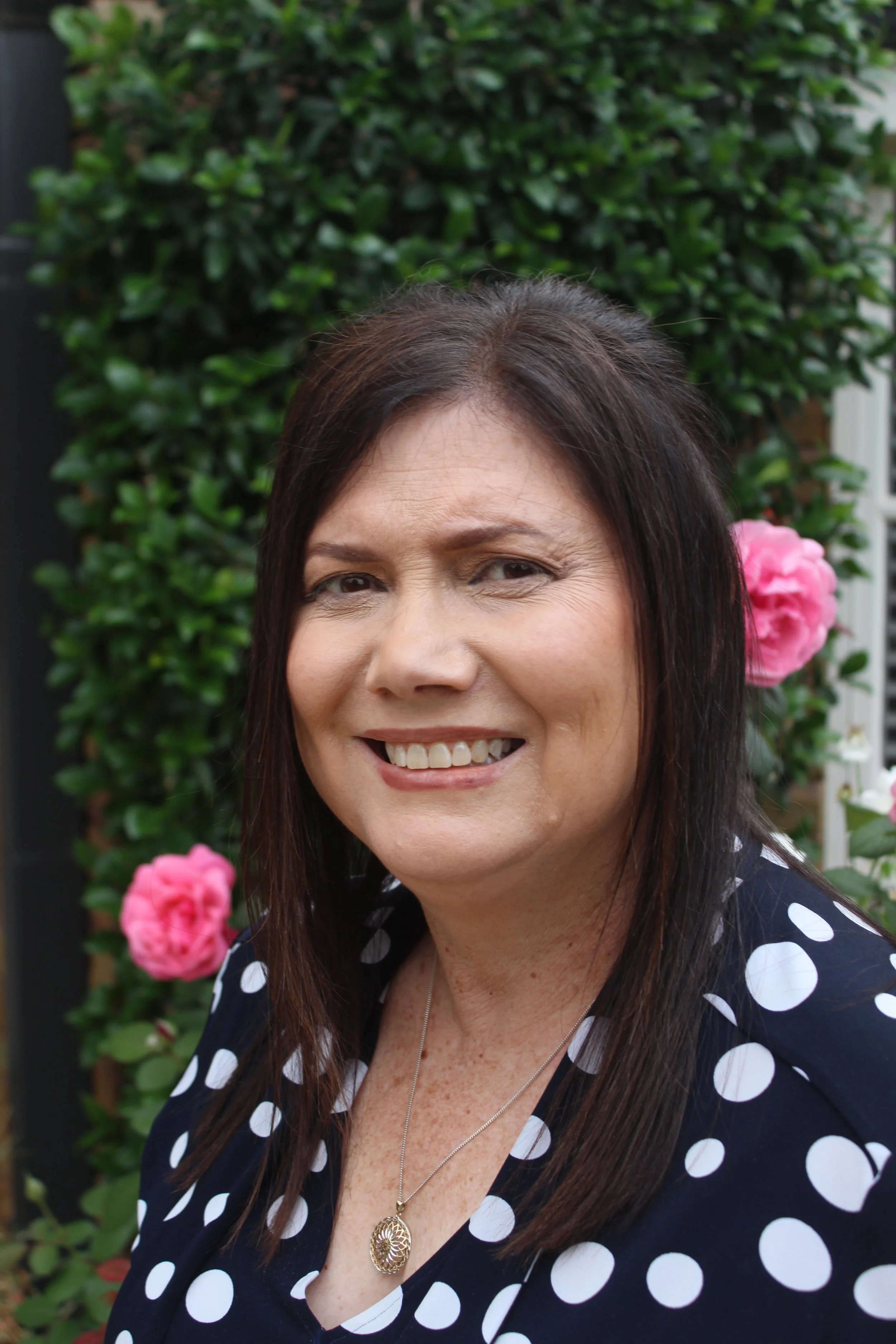 Portrait of a smiling woman with dark brown hair, wearing a navy blue polka dot top and a silver necklace with a pendant, standing outdoors in front of greenery and pink roses.