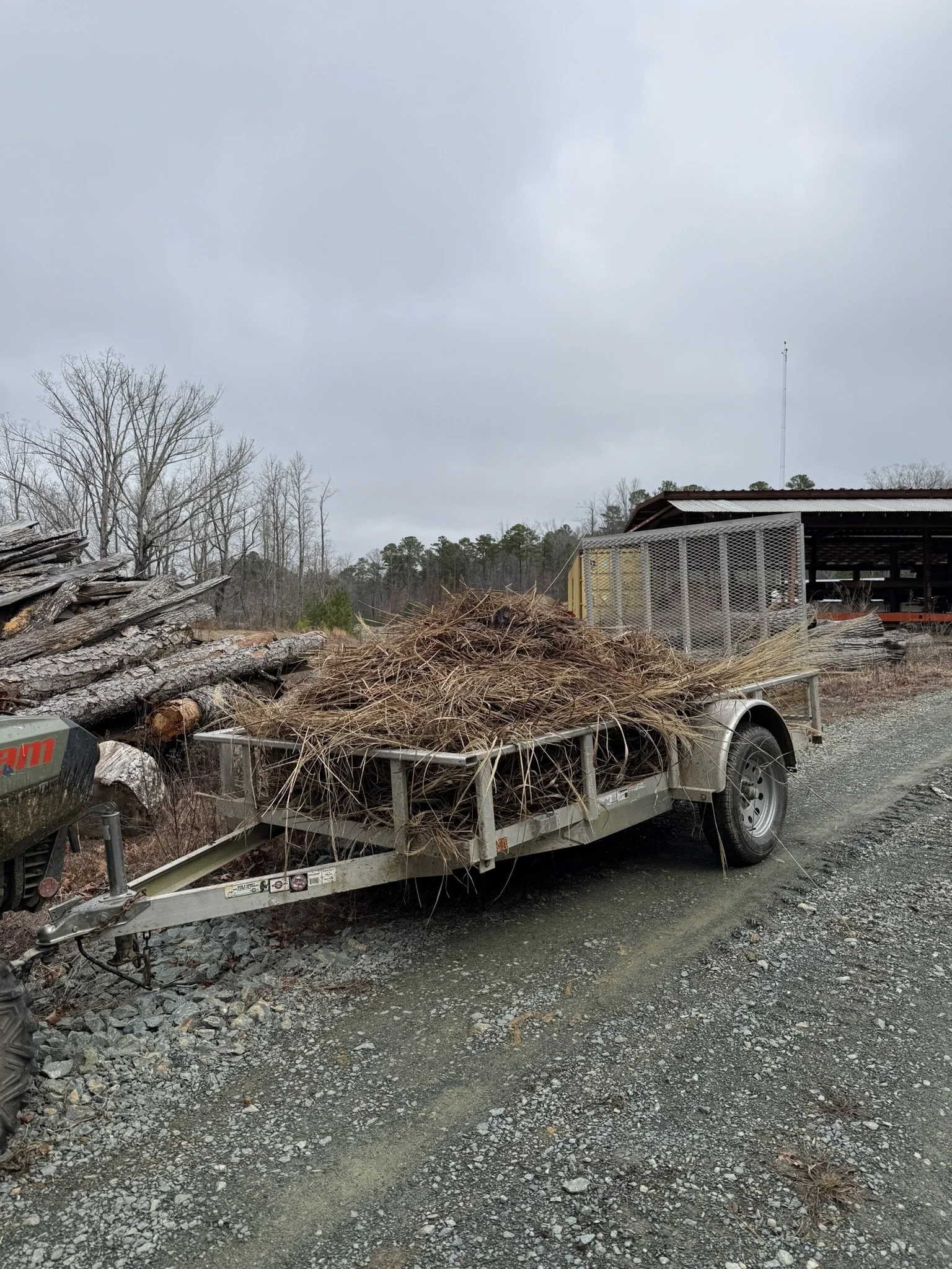 A trailer filled with dry grass and twigs parked on a gravel driveway with a cloudy sky above and trees in the background.