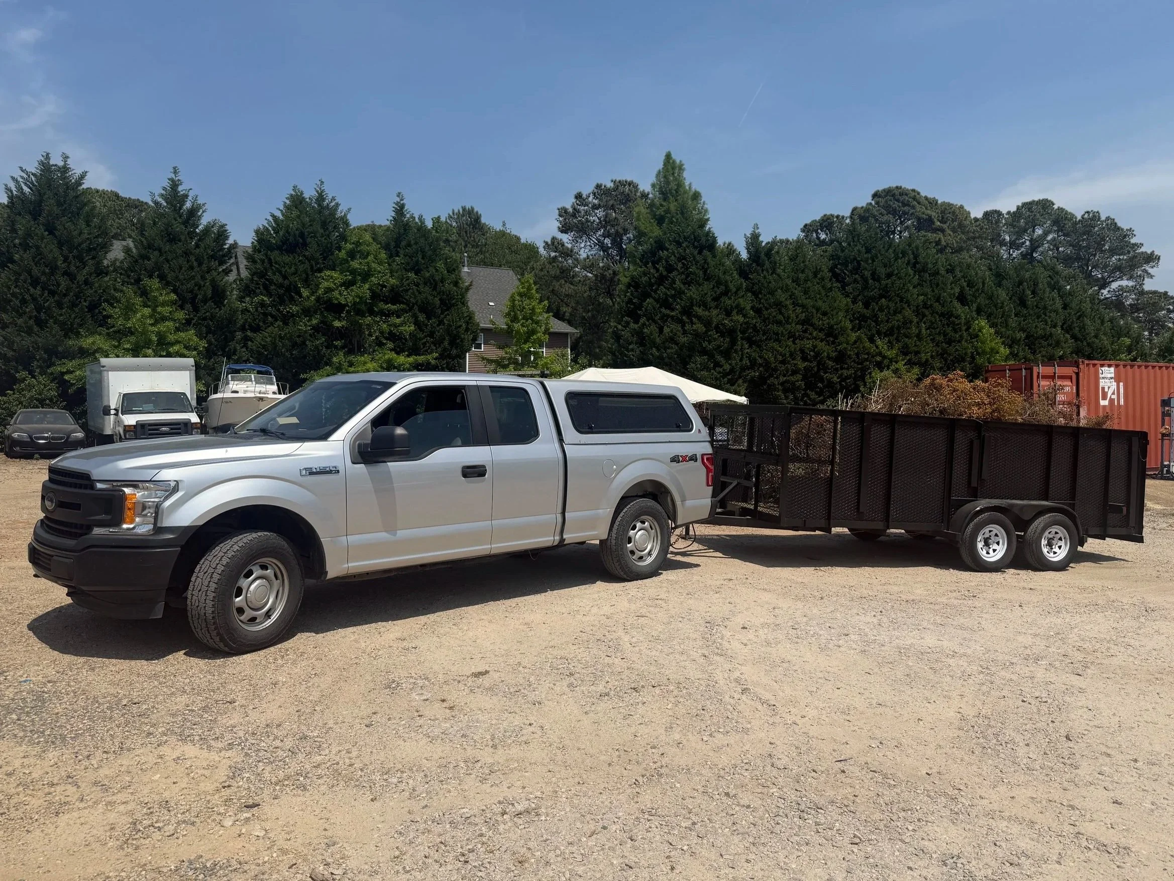 A white pickup truck attached to a black utility trailer parked on a gravel lot with trees and buildings in the background under a clear blue sky.