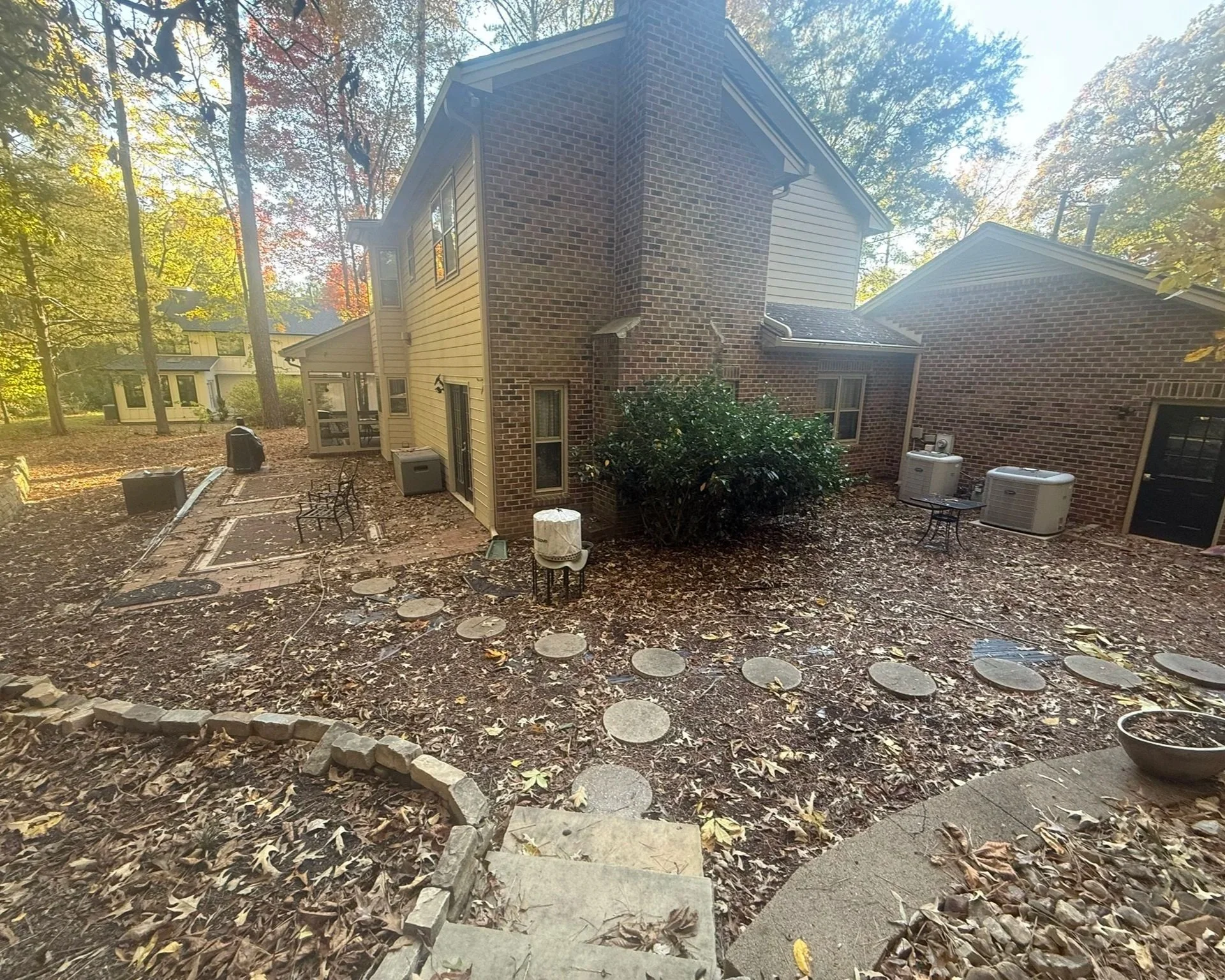 Backyard of a house with a brick and beige siding exterior, surrounded by fall leaves, trees, and outdoor air conditioning units, with a stone pathway and leaf-covered ground.