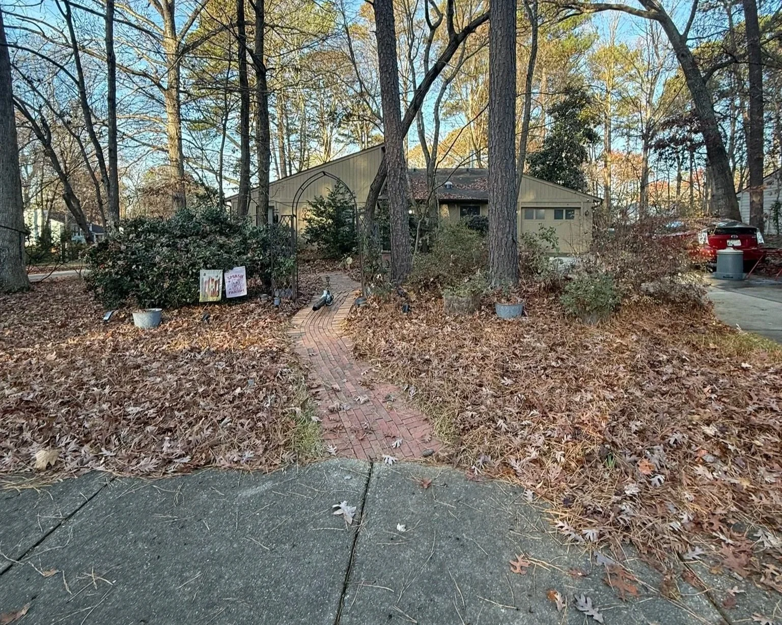 A front yard with leaf-covered ground, a curved brick walkway, and large trees. In the background, there is a house with a sloped roof, along with a car parked on the driveway and a trash bin. There are signs and pots near the walkway, and the sky is