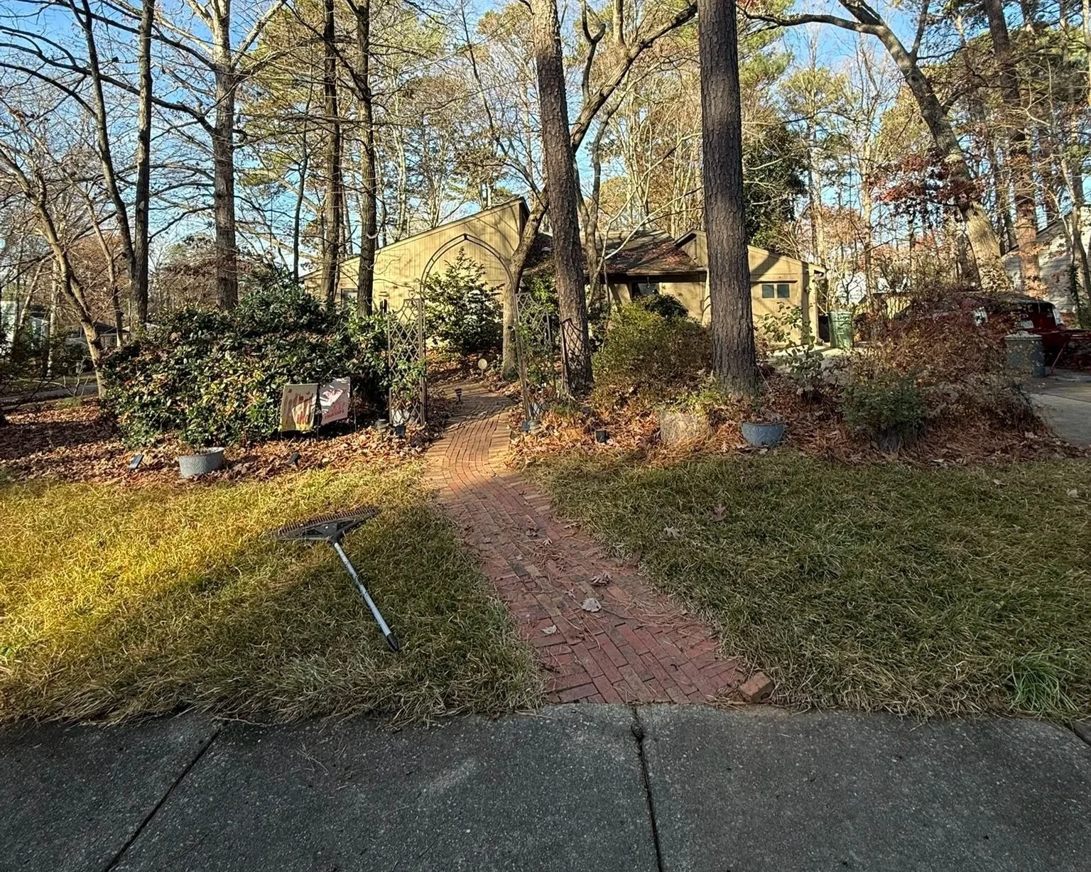 A curved brick pathway leading to a house surrounded by trees and bushes in a suburban yard, with a garden rake laying on the grass in the foreground.
