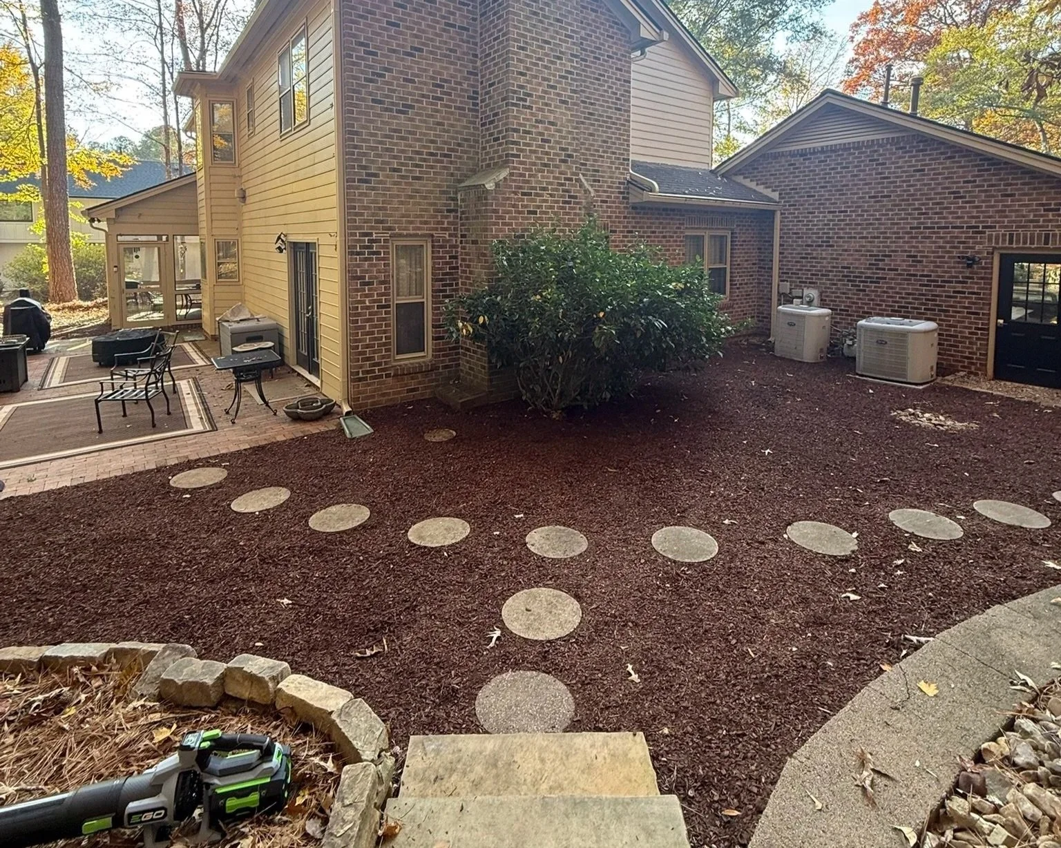 Backyard with newly installed circular stepping stones on reddish mulch, outdoor furniture on patio, bush, air conditioning units, and house with brick and siding exterior.