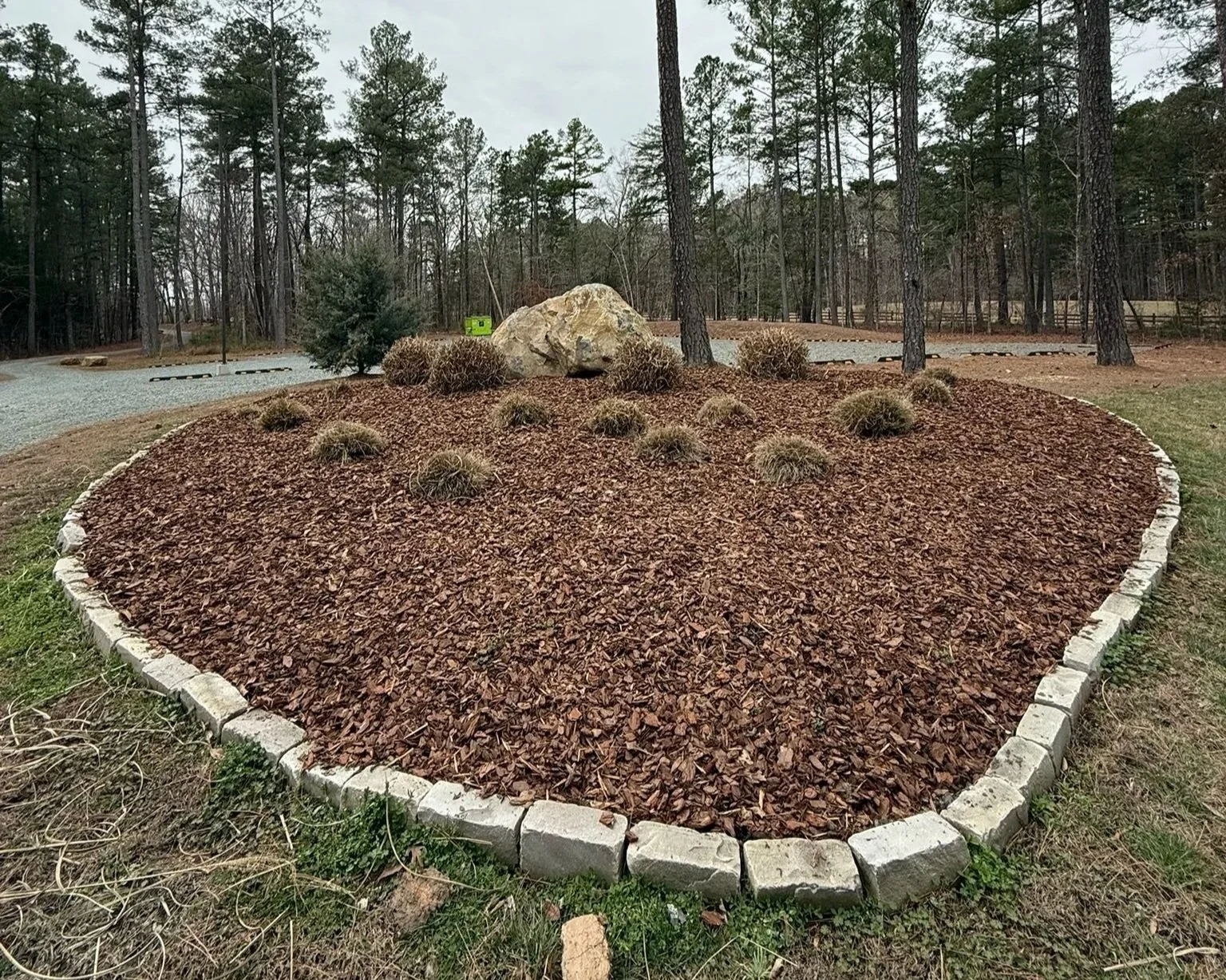 A landscaped garden bed with small bushes and a large rock, surrounded by a border of light-colored stones, in a park or wooded area with tall trees in the background.