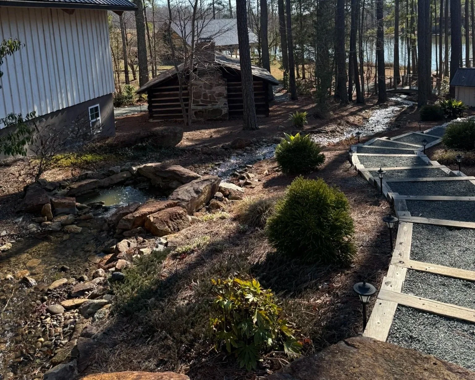 A landscaped backyard with a small stream with rocks, a stone and wood shed, a winding gravel path, bushes, trees, and outdoor lights.