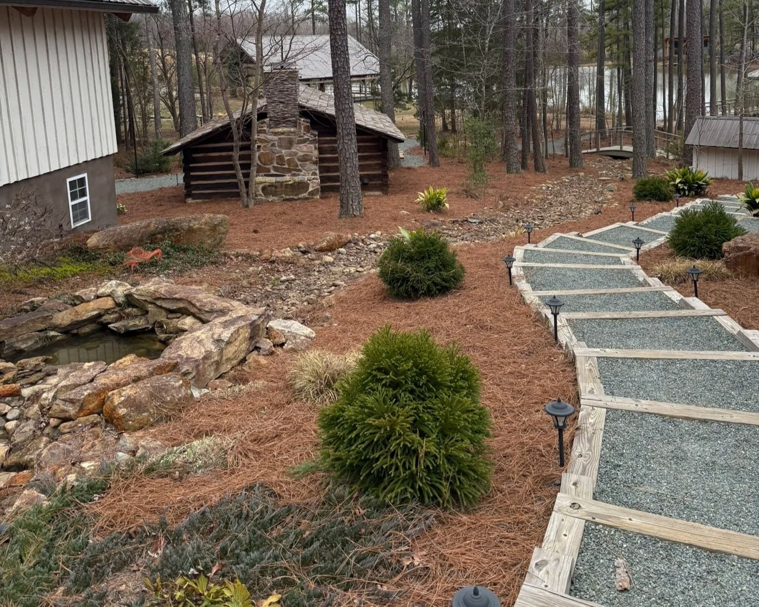 A landscaped backyard with a stone and gravel pathway, small bushes, pine trees, a pond with rocks, and a small wooden cabin in the woods.