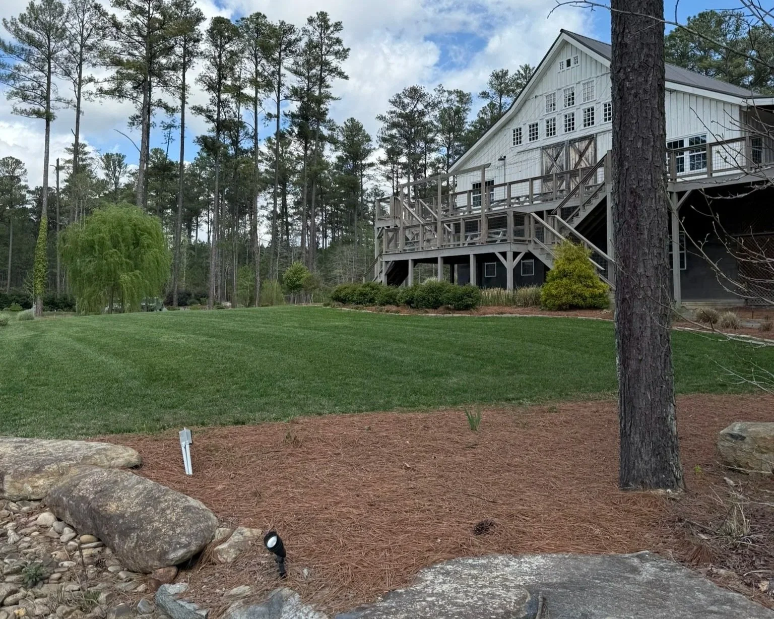 A commercial venue property with a deck and stairs, surrounded by recently mowed green grass, trees, and landscaping under a cloudy sky in Raleigh.