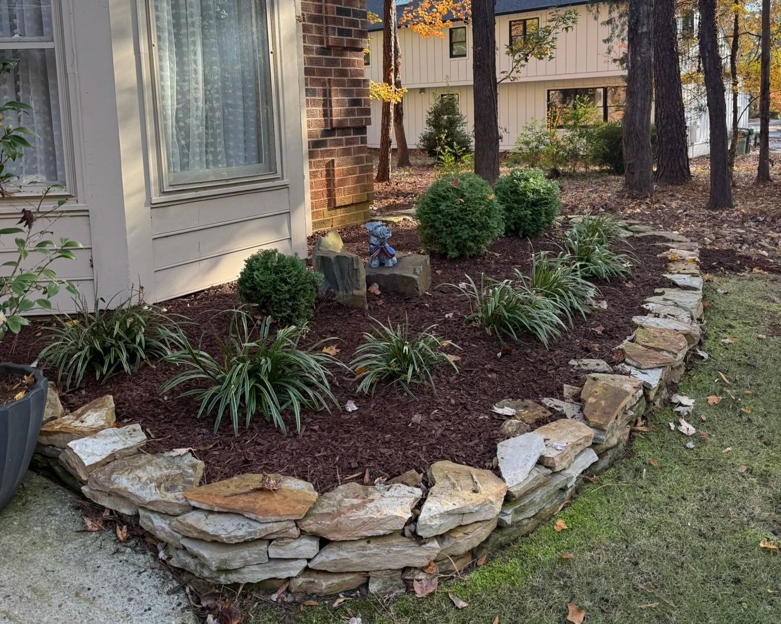 A landscaped garden bed with small shrubs and ornamental grass, edged with a low stone wall, next to a house with a brick and siding exterior, and surrounded by trees with fall foliage.