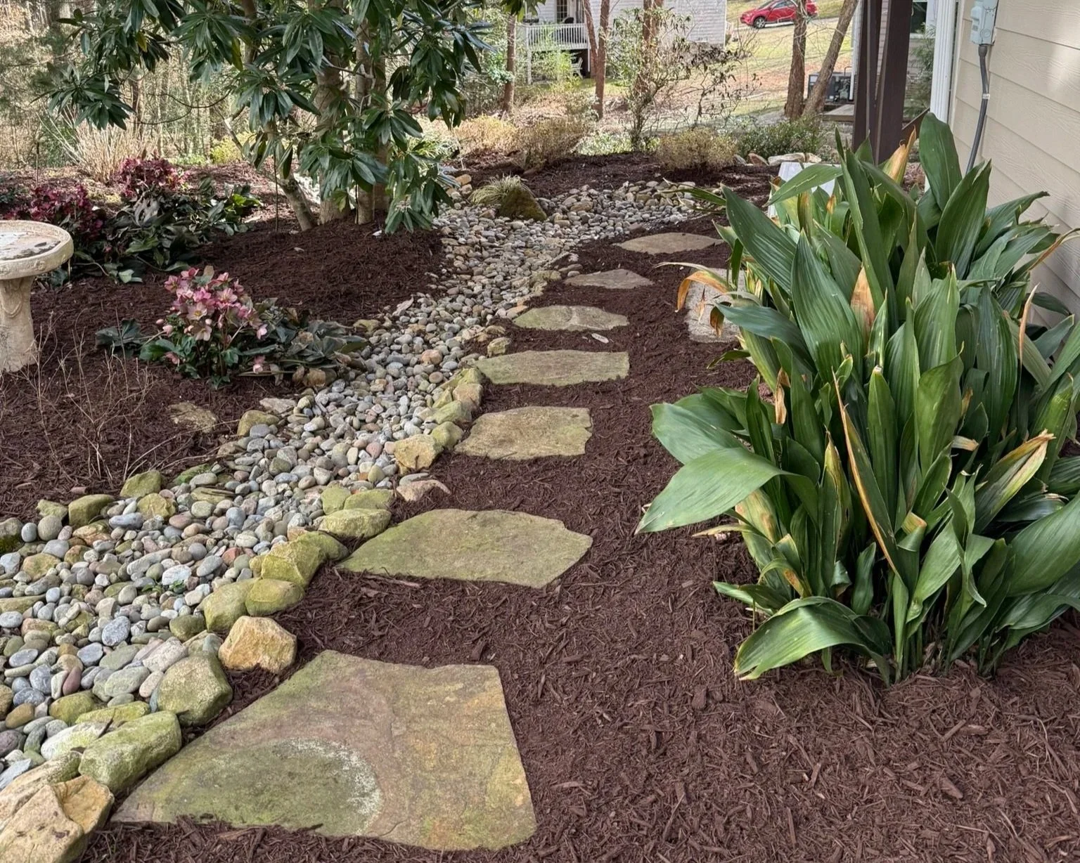 A garden pathway made of large, flat stepping stones bordered by small, rounded rocks and mulch, surrounded by green shrubs and plants.