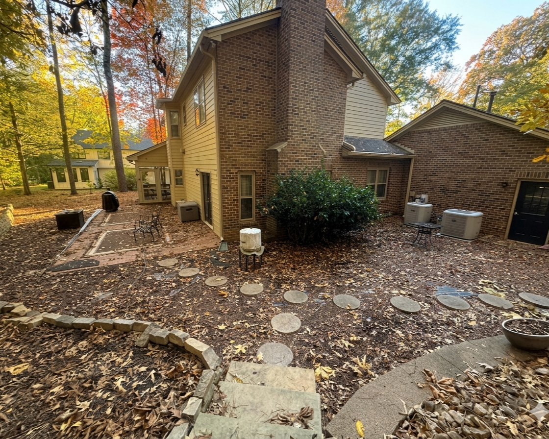 Backyard patio area of a house with fallen autumn leaves on the ground, round stepping stones creating a pathway, and trees with changing fall foliage in the background.
