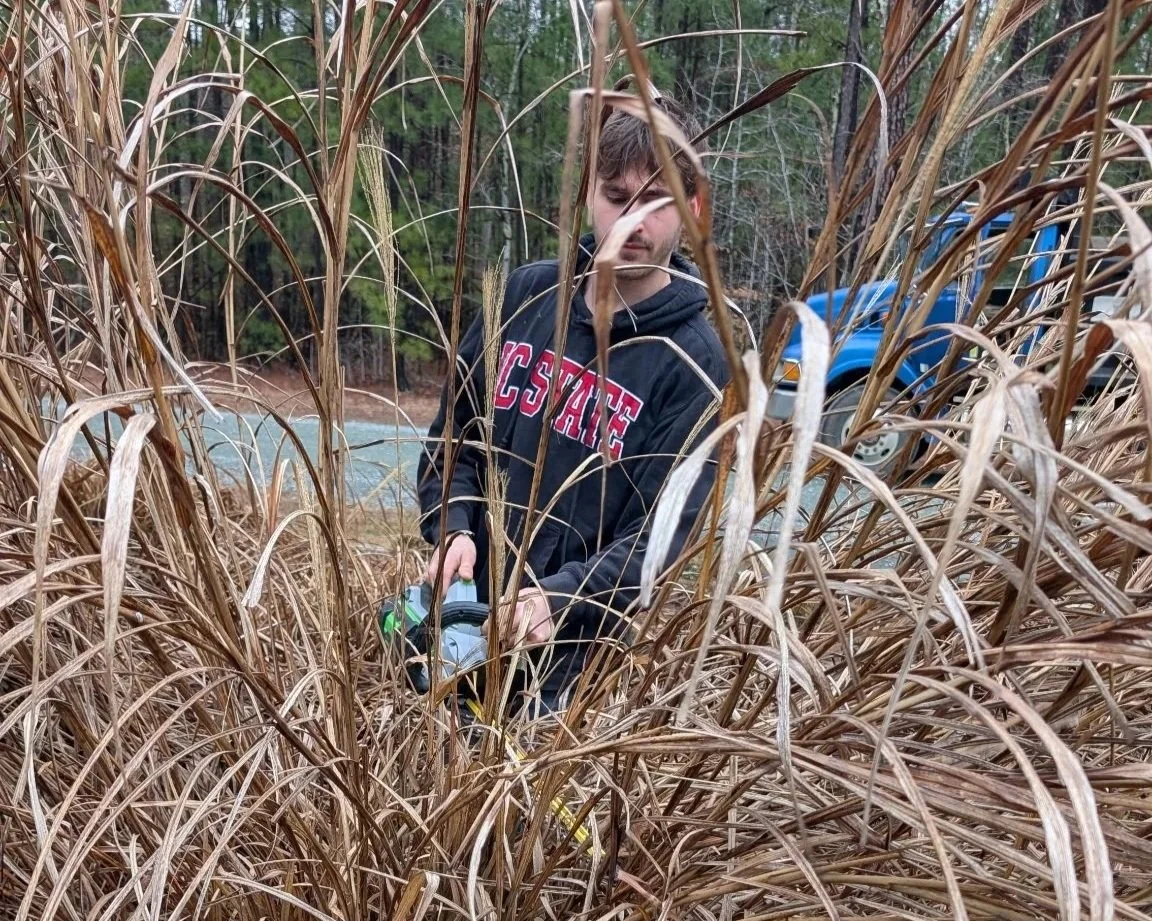 A young man in a black hoodie with 'CSU' written on it is standing among tall, dry grass near a body of water and a parked blue truck, holding a device resembling a small gas-powered tool, possibly a weed trimmer.
