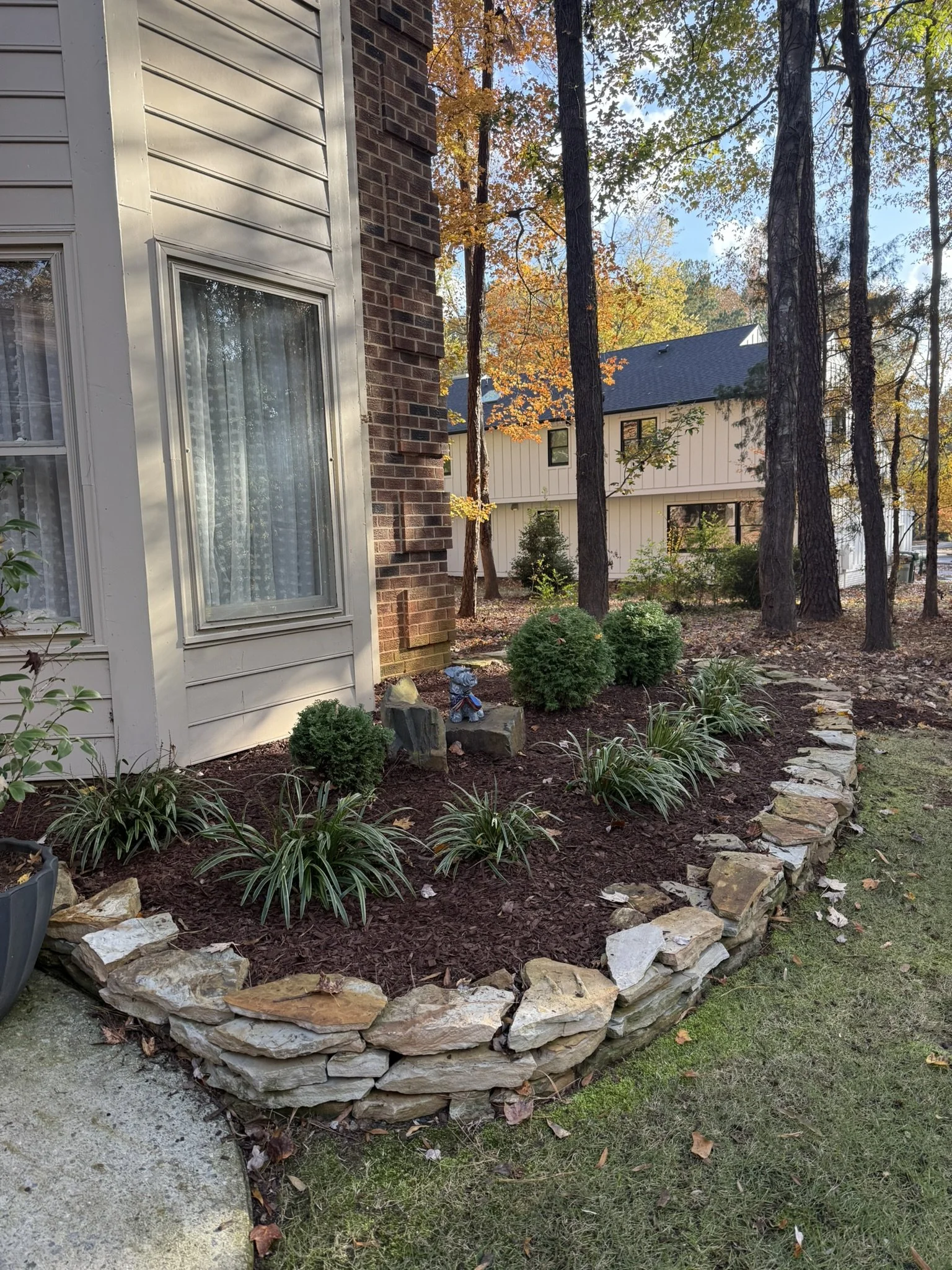 A landscaped garden bed with small shrubs and plants, bordered by stacked rocks, next to a house with brick and siding exterior, in a backyard with trees and leaves on the ground.