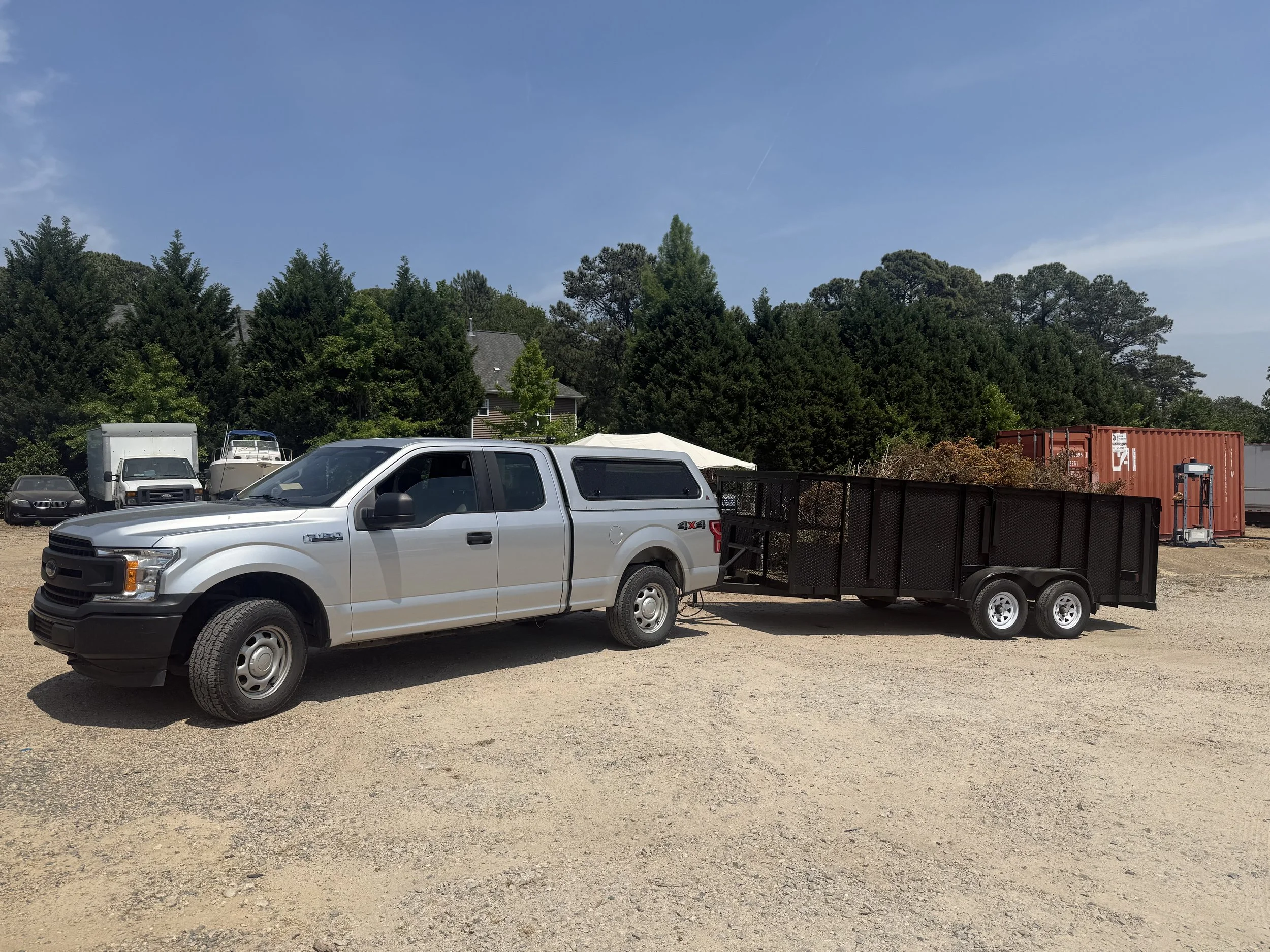 A parking lot with a white pickup truck attached to a black trailer, several other vehicles including a white van and a boat, a red shipping container, a fence, trees, and a house with a gray roof under a blue sky with some clouds.