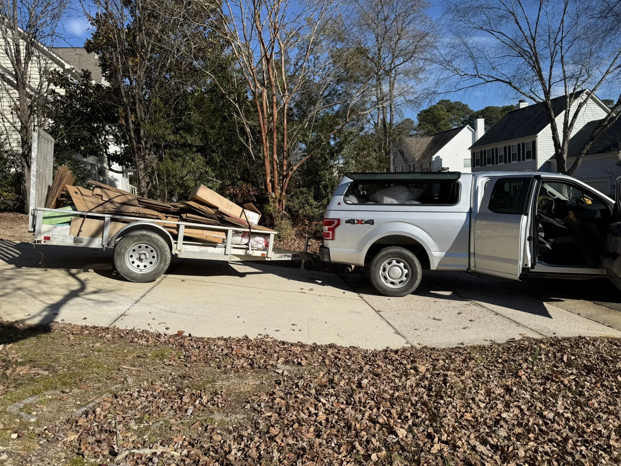 A white pickup truck attached to a trailer carrying cardboard and wood debris parked on a driveway in a neighborhood with trees and white houses.