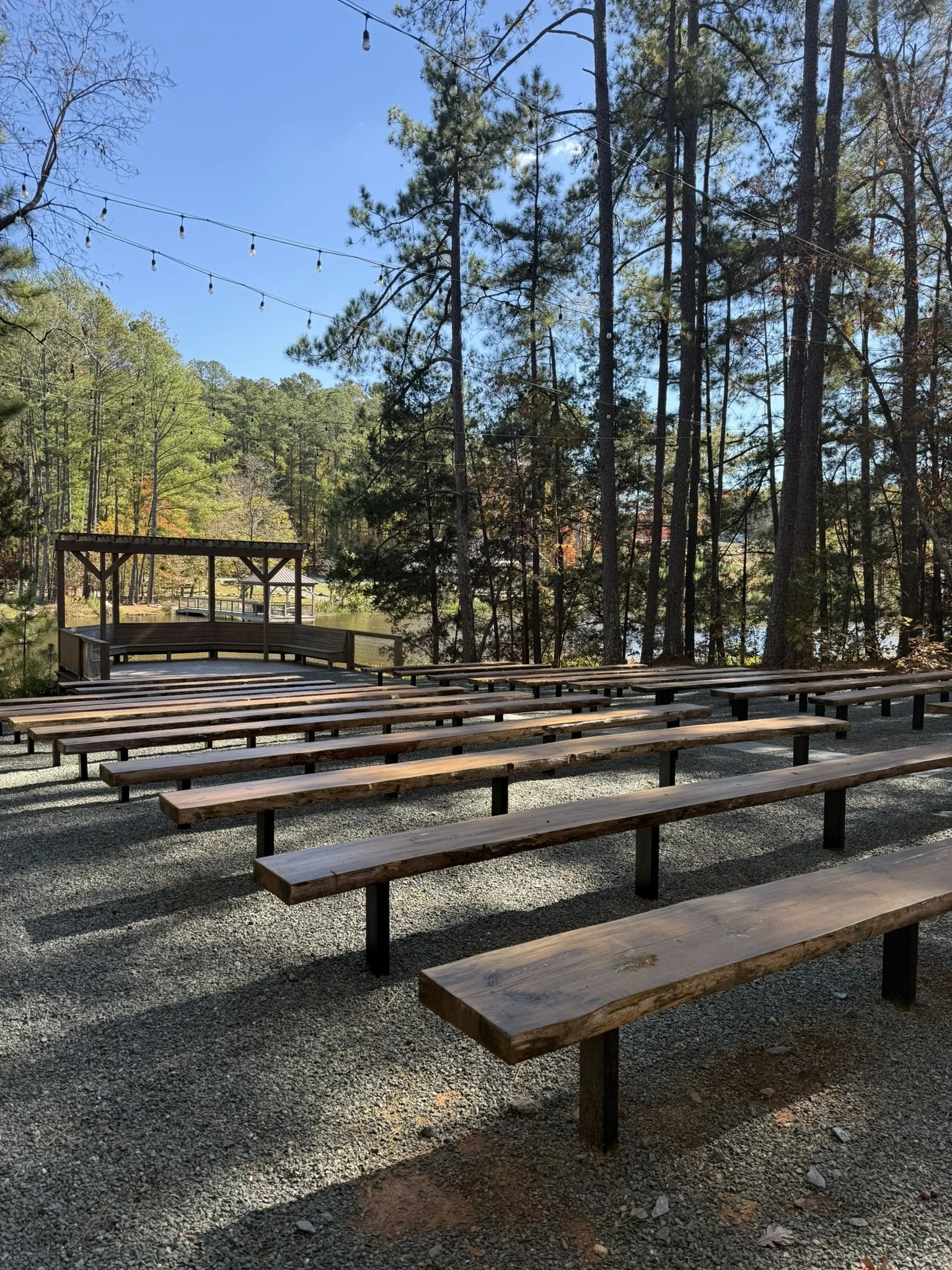 Outdoor amphitheater with wooden benches on gravel, surrounded by trees, with string lights hanging overhead on a clear day.