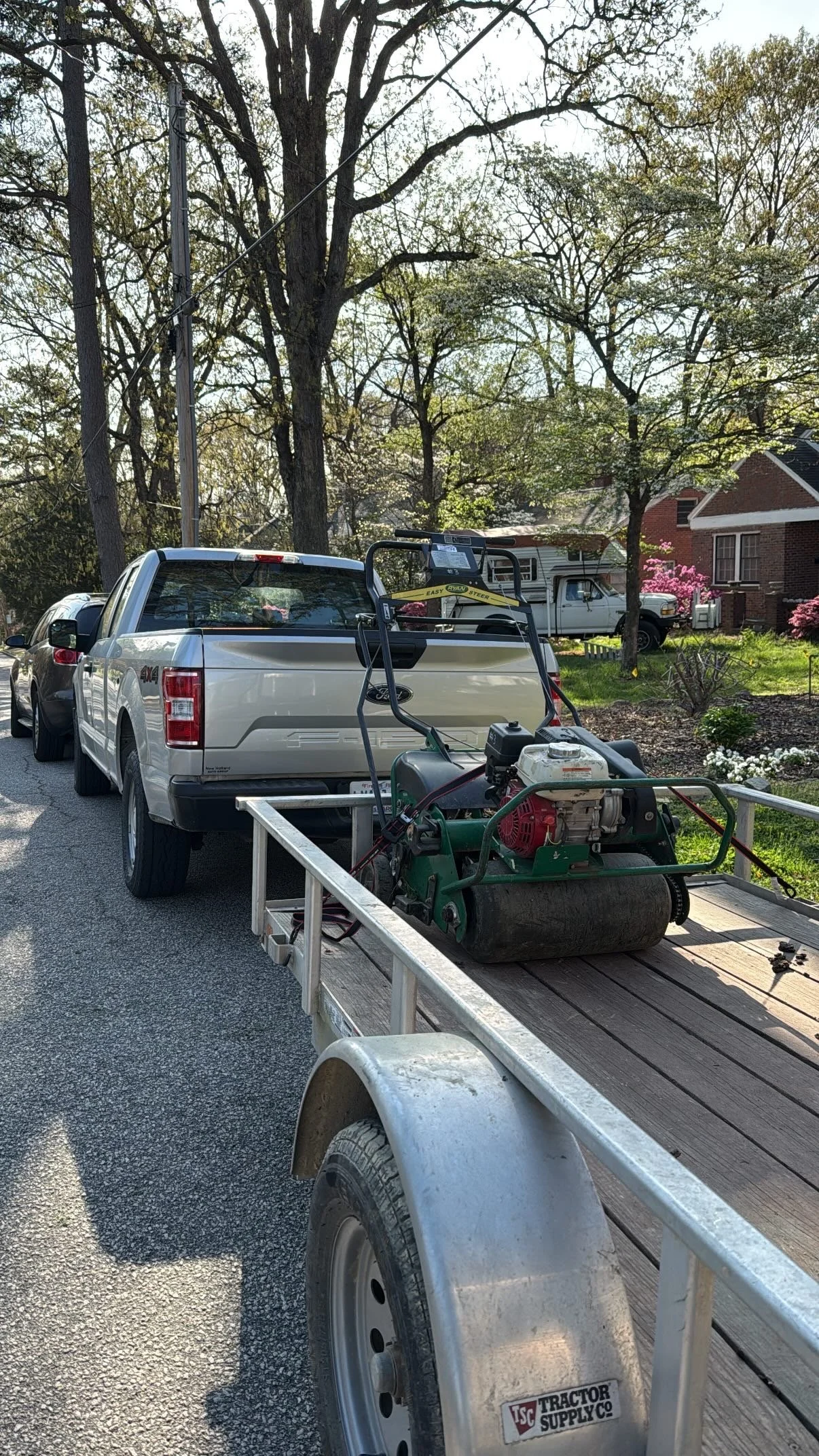 A flatbed trailer attached to a white Ford pickup truck, with a large lawn roller on the trailer, parked on a residential street with trees and houses in the background.