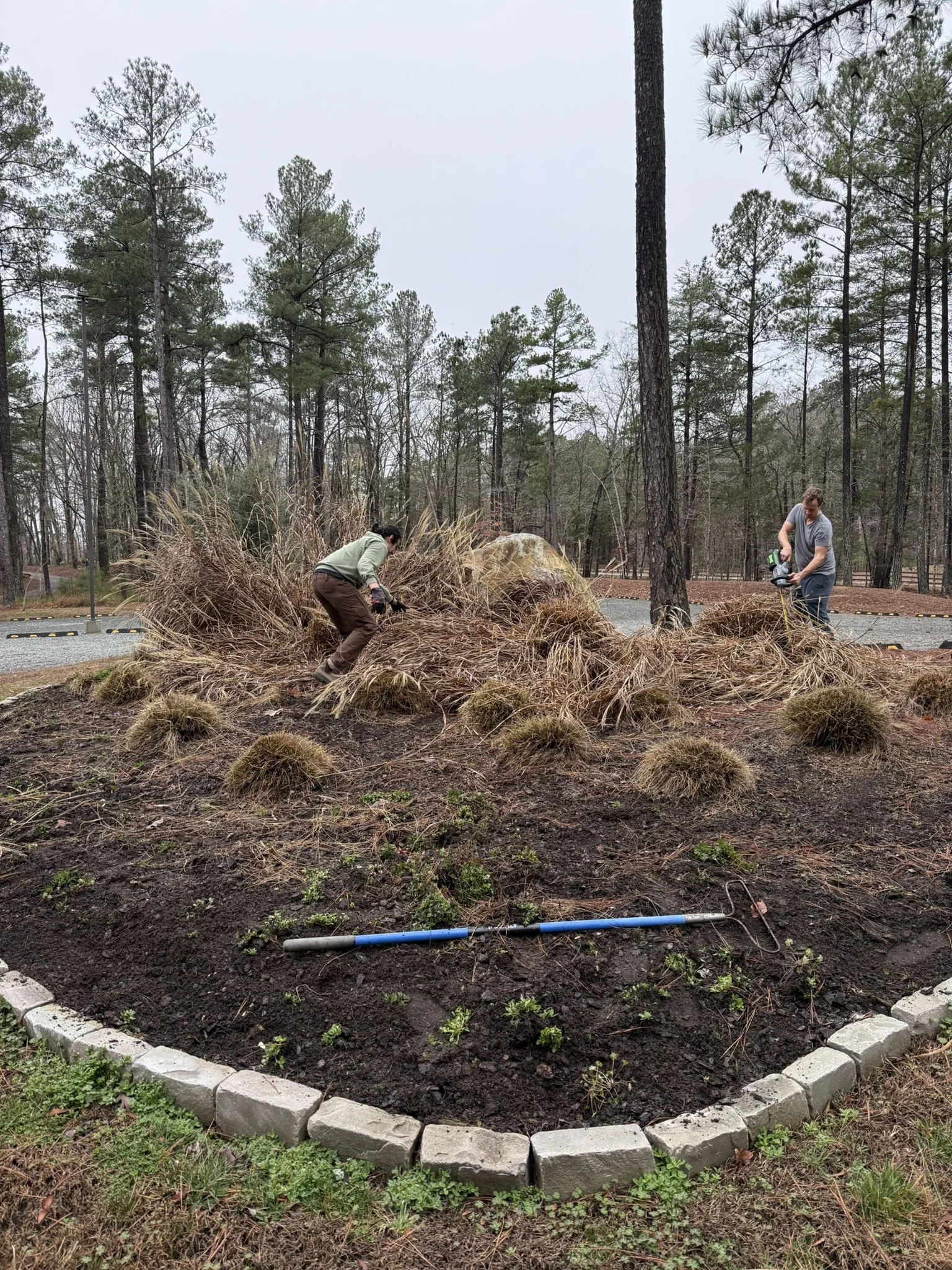 Two people are working on a garden bed surrounded by a border of gray stones. One is using a gardening tool, and the other is working with landscaping material, with a wooded area in the background.