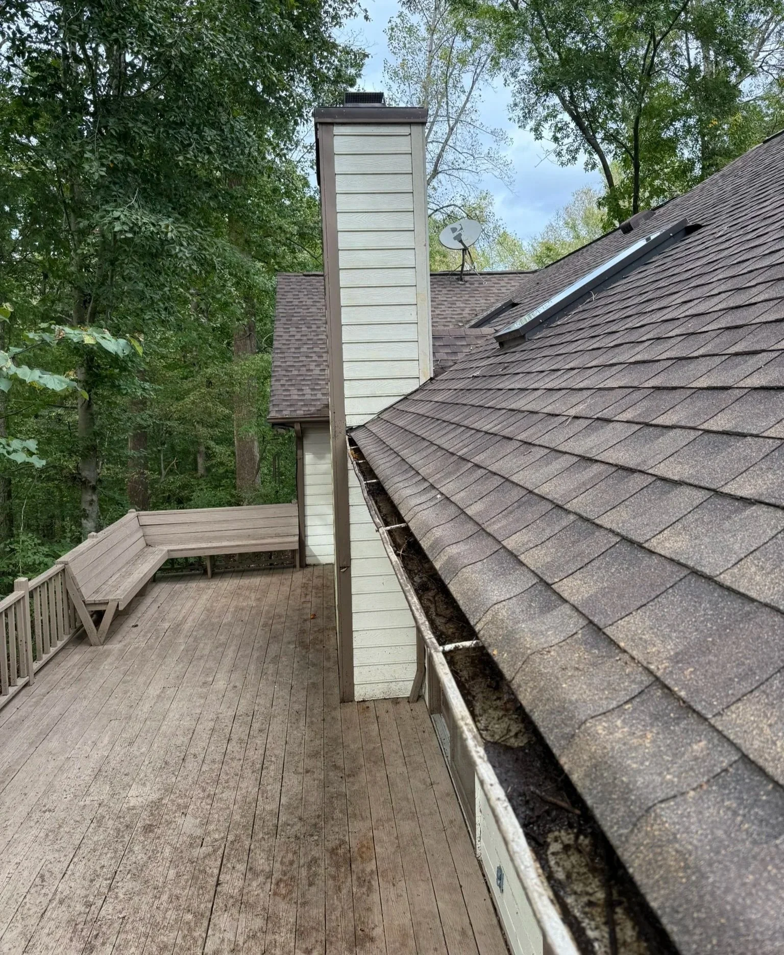 A backyard deck with a wooden railing, a built-in bench, and a house with a steeply pitched roof, chimney, and satellite dish surrounded by trees.