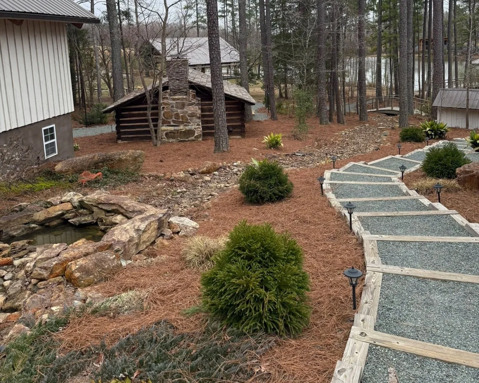 Backyard garden with a stone pathway, small shrubs, a pond with rocks, tall trees, and a rustic log cabin with a stone chimney in the background.