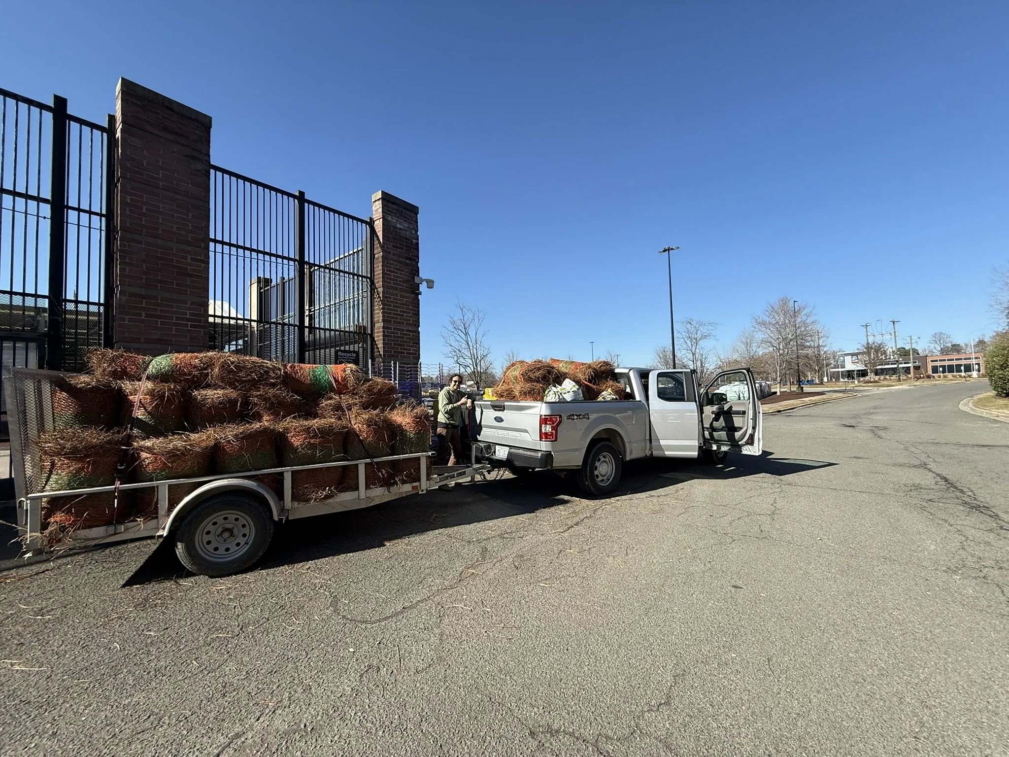 A white pickup truck with an open tailgate and two open doors parked in a parking lot. The truck is loaded with rolled up straw or hay bales and some bags. There is a person standing beside the truck, holding a piece of straw or hay. The background features a brick fence with black metal railings and a clear blue sky.