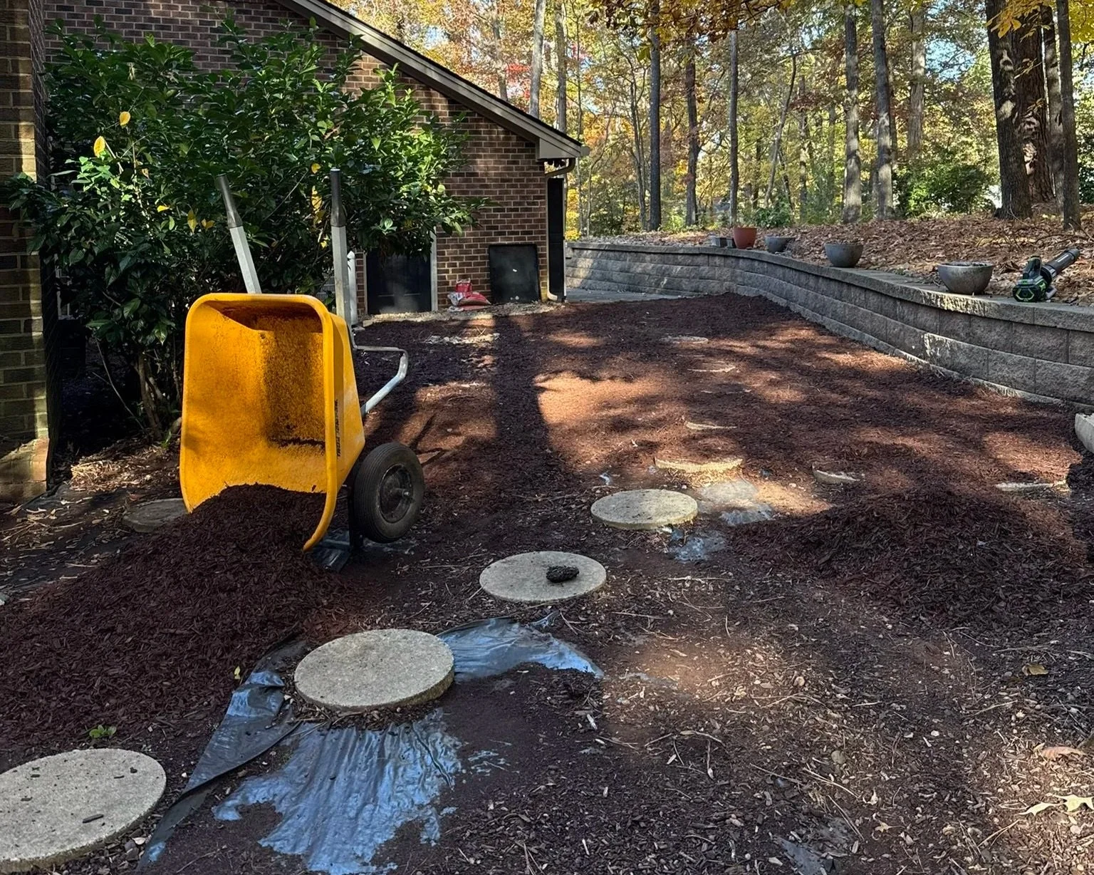 A backyard area under landscaping renovation with a yellow wheelbarrow tipped over and soil spilling out, circular stepping stones laid out, a brick house on the left, a retaining wall with potted plants on top, and trees in the background.