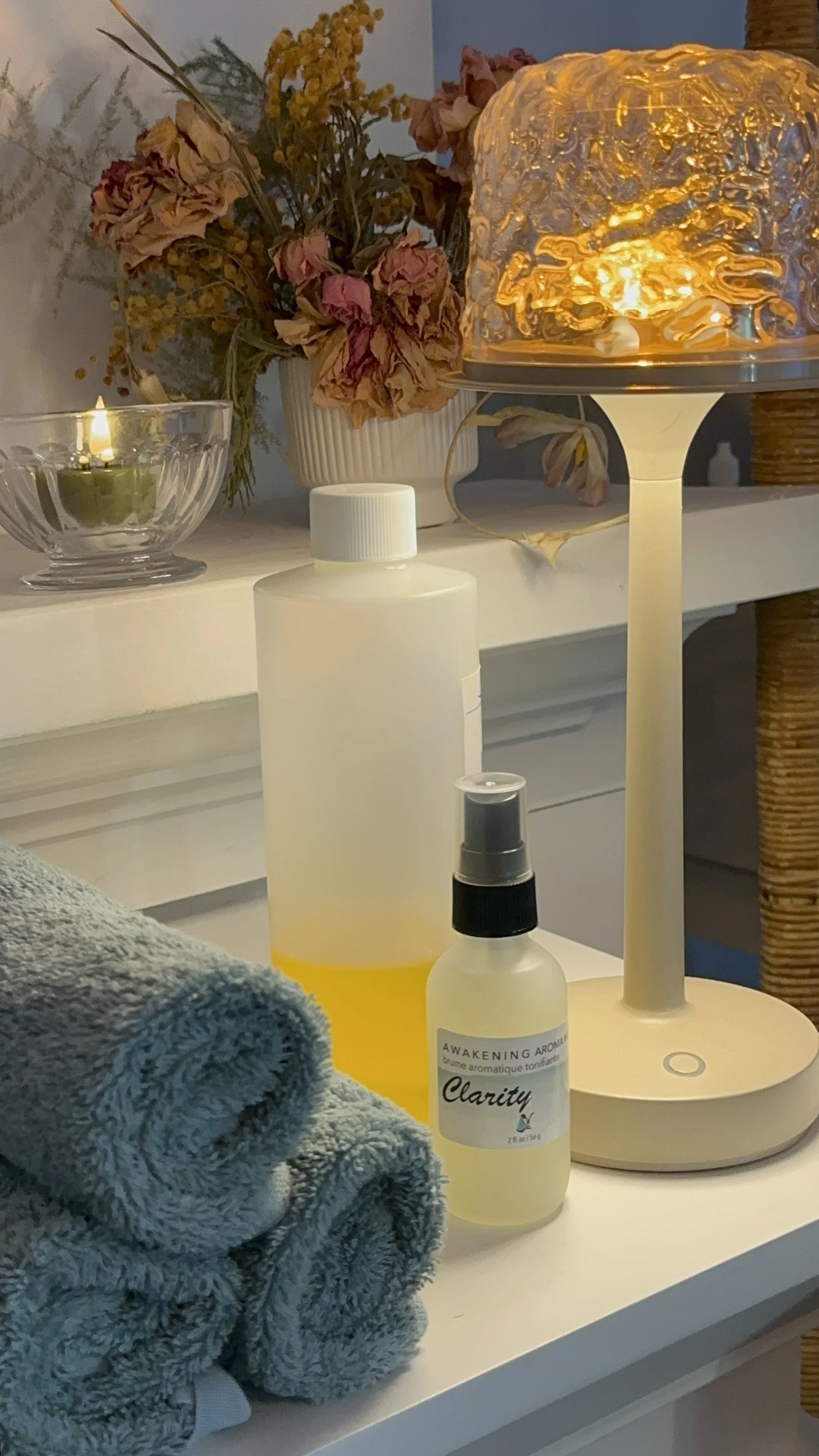 Close-up of a white surface with a rolled grey towel in the foreground. On the surface, there are several skincare or aromatherapy bottles, a diffuser emitting a warm glow, a small glass bowl with a lit candle, and a flower arrangement with dried pink and yellow flowers in a white vase.
