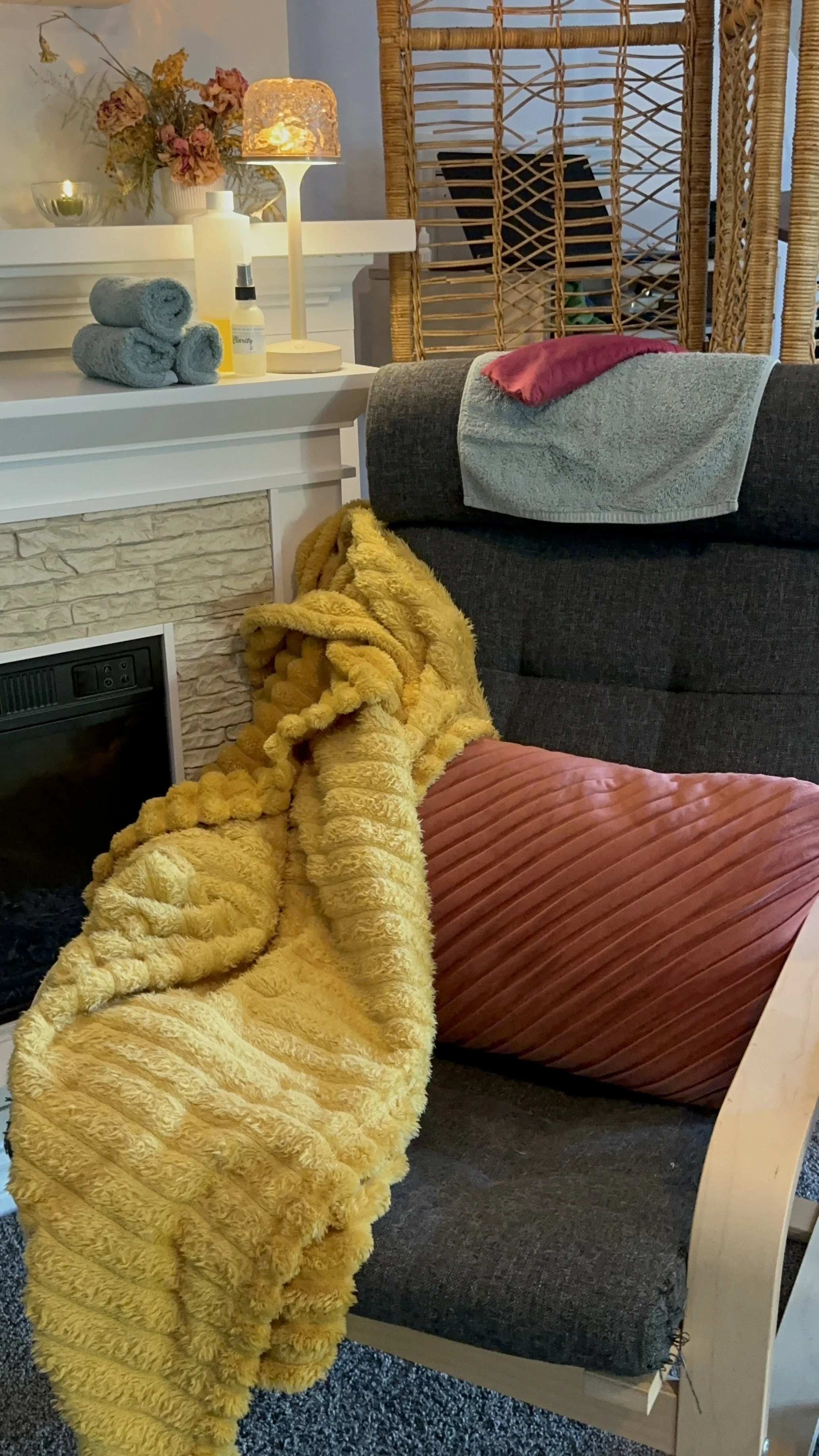 A cozy living room corner with a dark gray sofa, a yellow plush blanket, a pink pillow, and a light-colored wooden furniture piece with towels, a lamp, and decorative items on top, with a wicker room divider and a blue wall in the background.