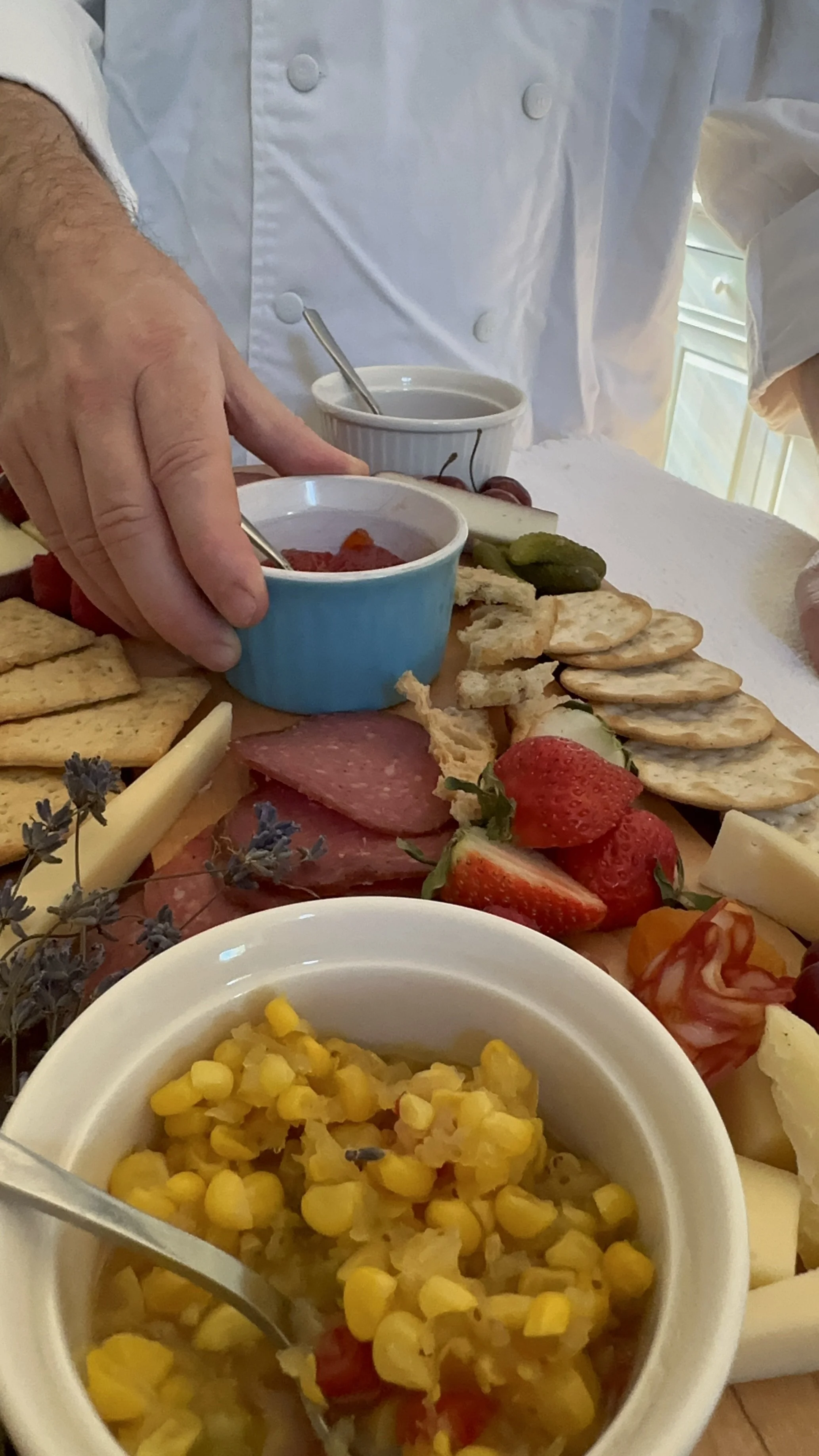 Close-up of a cheese and charcuterie platter with strawberries, crackers, pickles, and various cheeses, accompanied by bowls of condiments and corn salad, with a person in a chef's coat in the background.