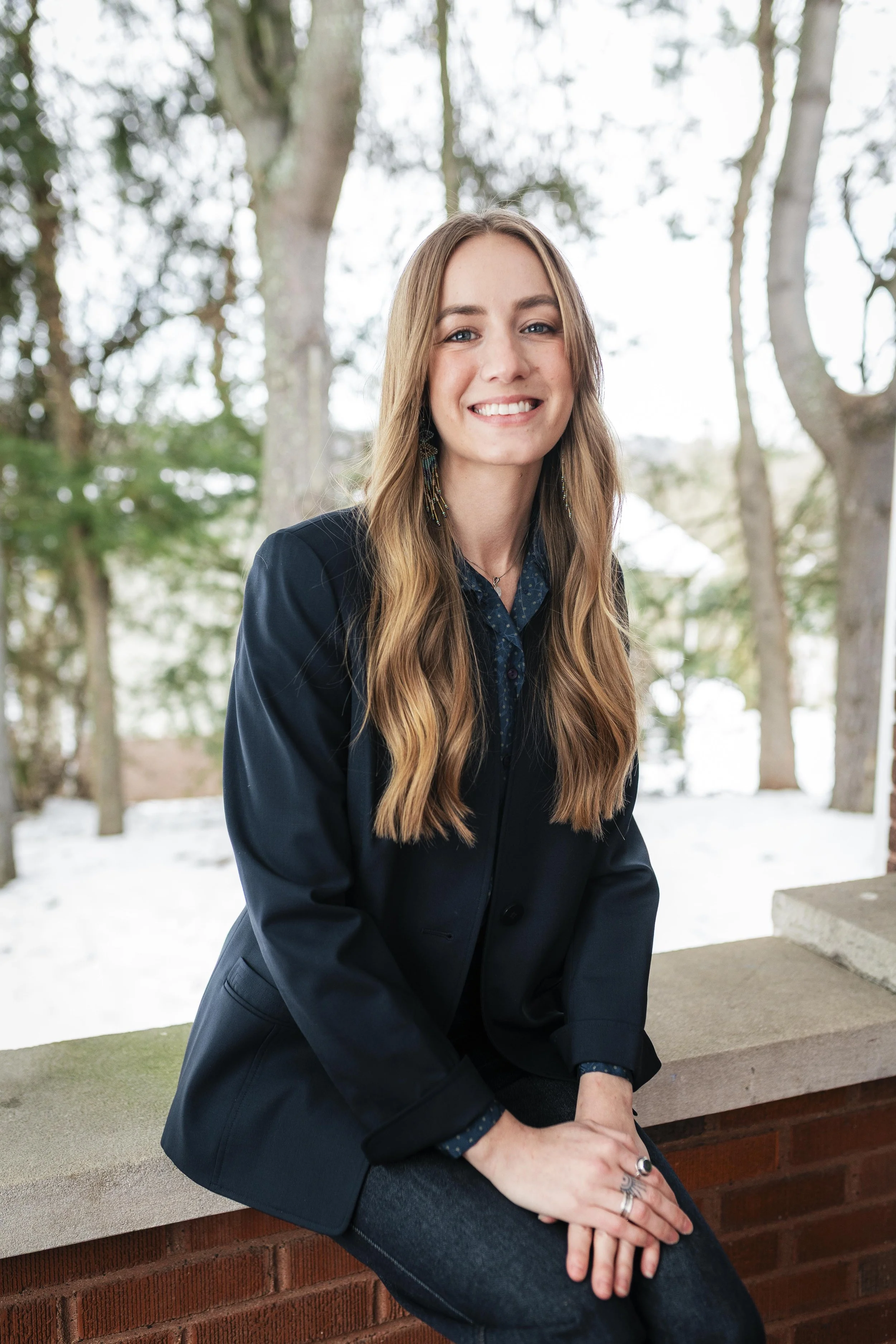 Smiling woman with long wavy hair in a black jacket, sitting on a brick ledge outdoors surrounded by trees and snow.