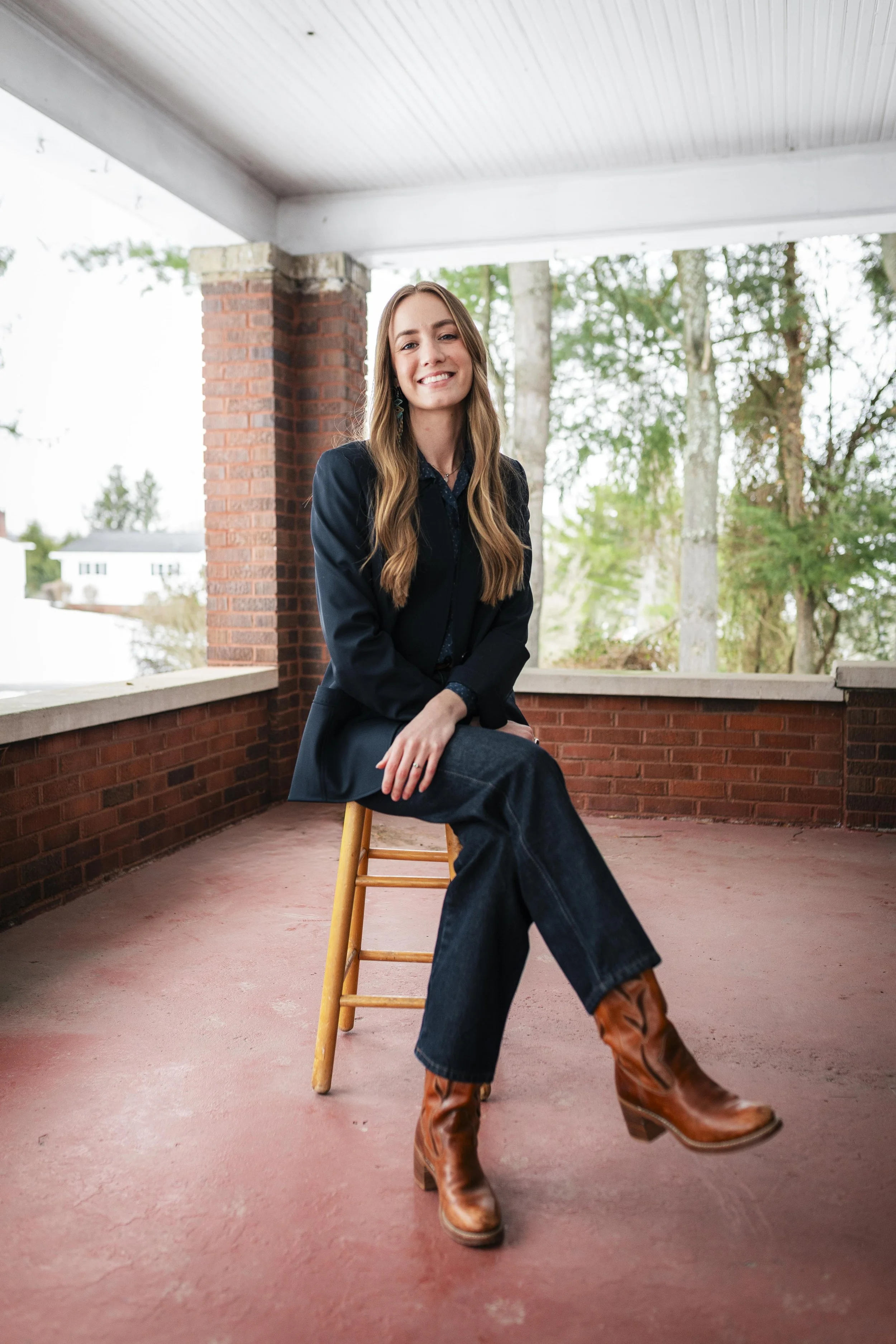 A young woman sitting on a wooden stool on a covered porch, smiling, wearing a black blazer, dark jeans, and brown cowboy boots, with trees and houses in the background.