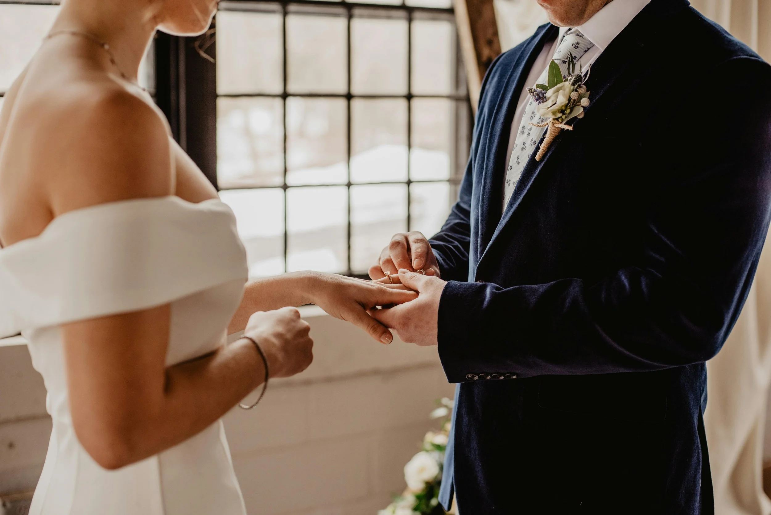 A couple exchanging wedding rings during their ceremony, with the bride in an off-the-shoulder white gown and the groom in a dark suit, near a window with natural light.