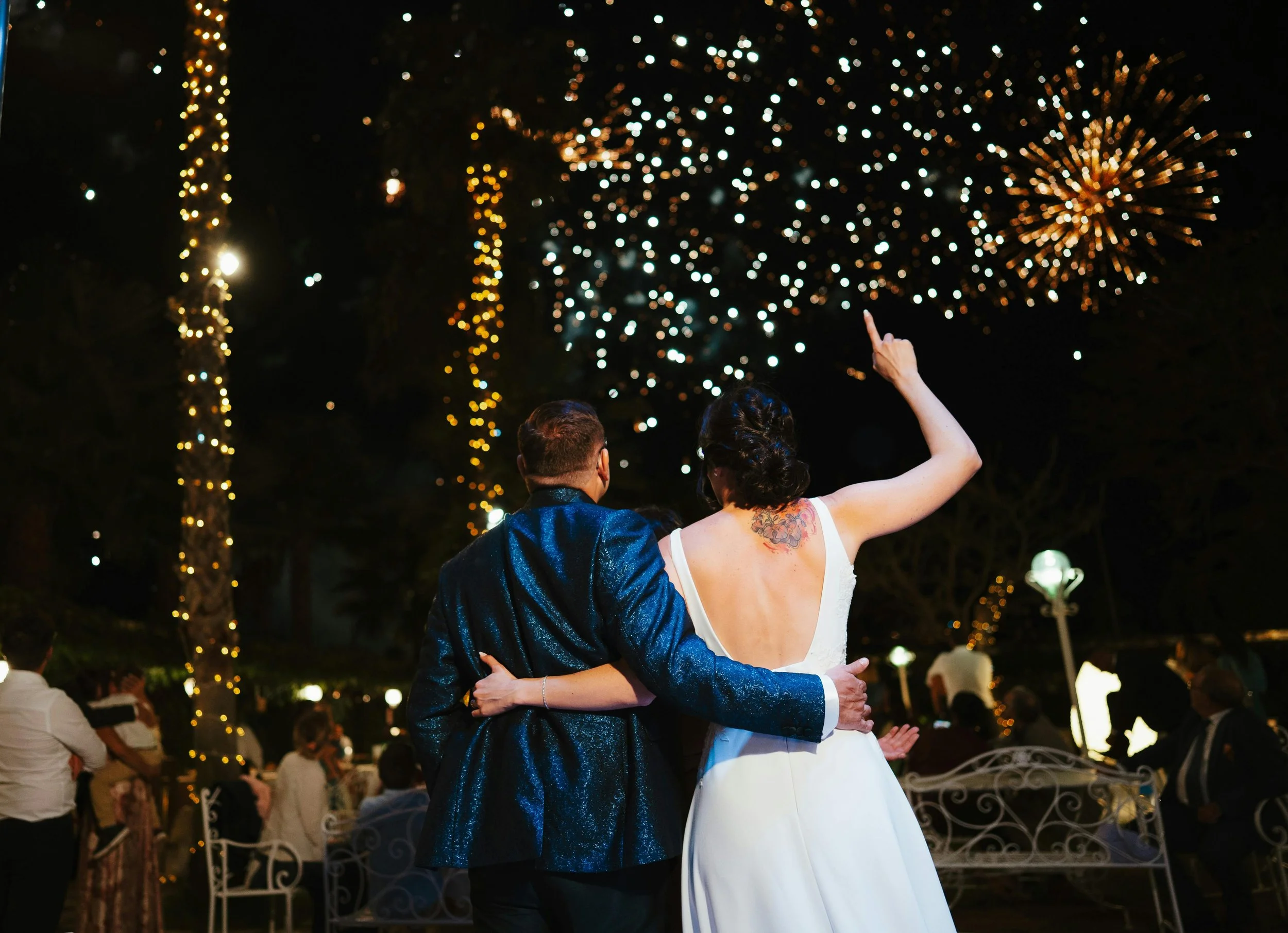 Couple celebrating at night under fireworks with string lights and trees in the background