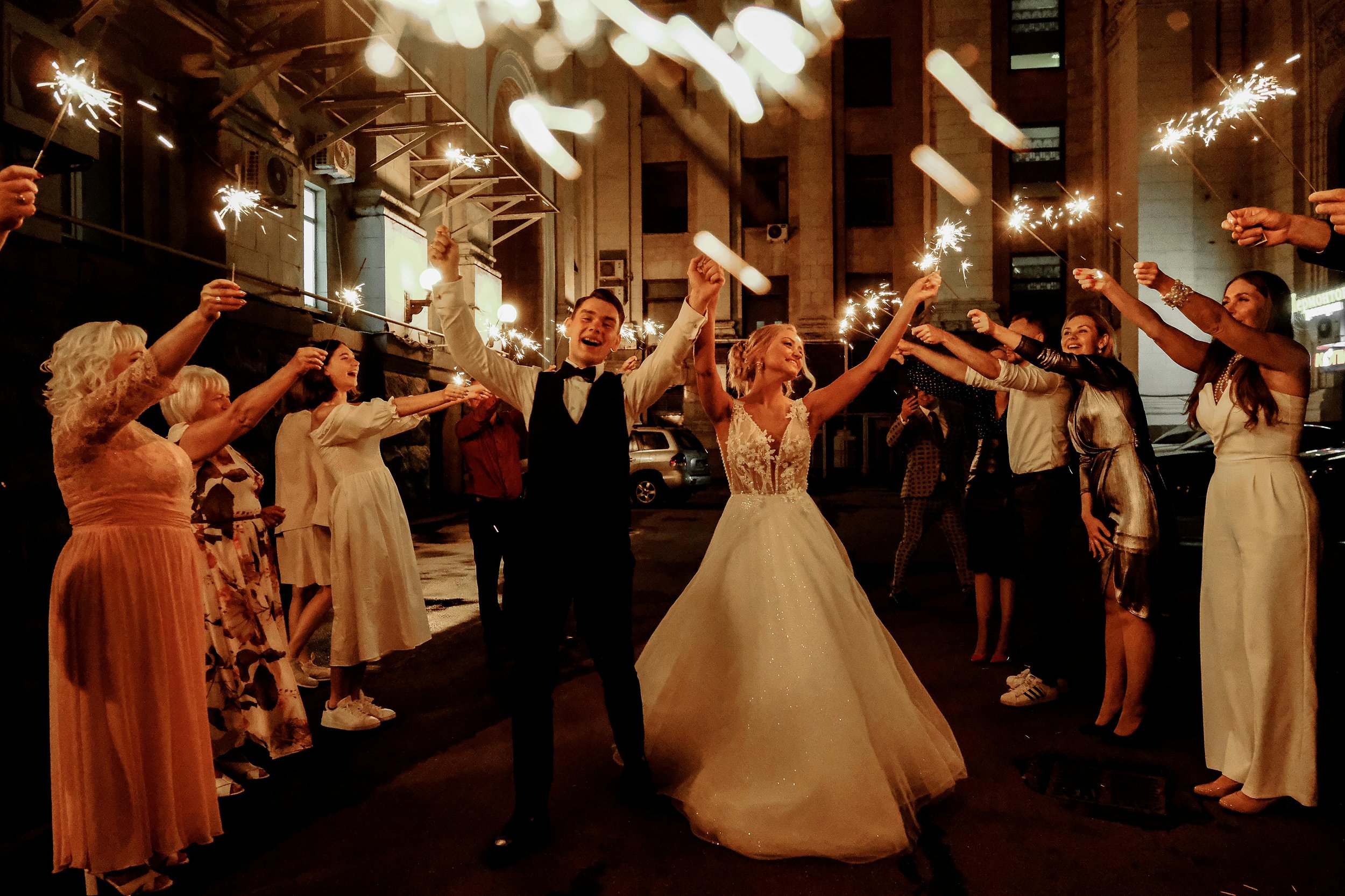 A couple dressed in wedding attire celebrating with sparklers surrounded by friends and family at night in an urban setting.