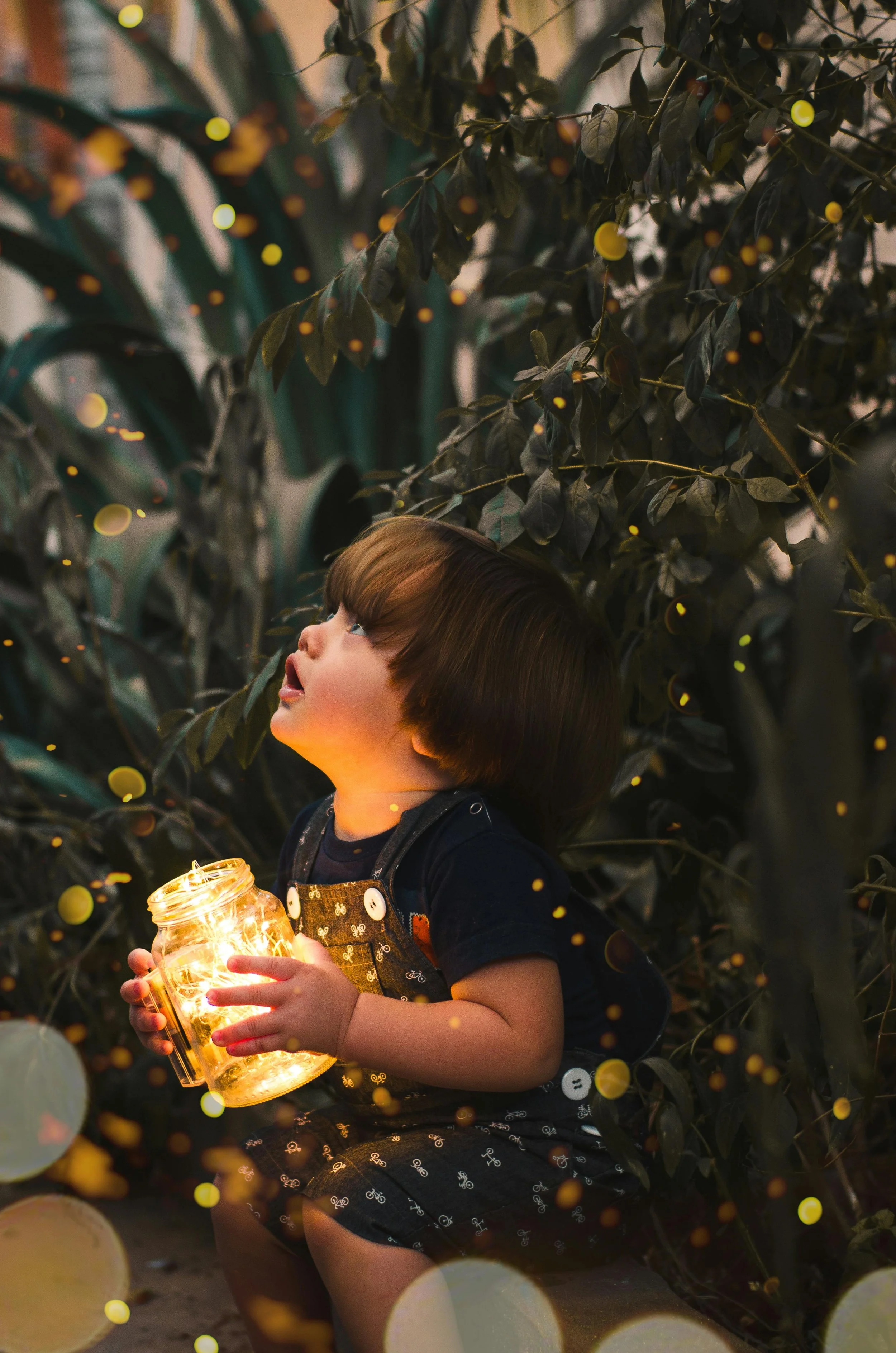 A young child with brown hair holding a jar of fairy lights, sitting amid dark green leafy plants, with a curious expression as they look upward. The scene is illuminated by the warm glow of the lights, creating a magical atmosphere.