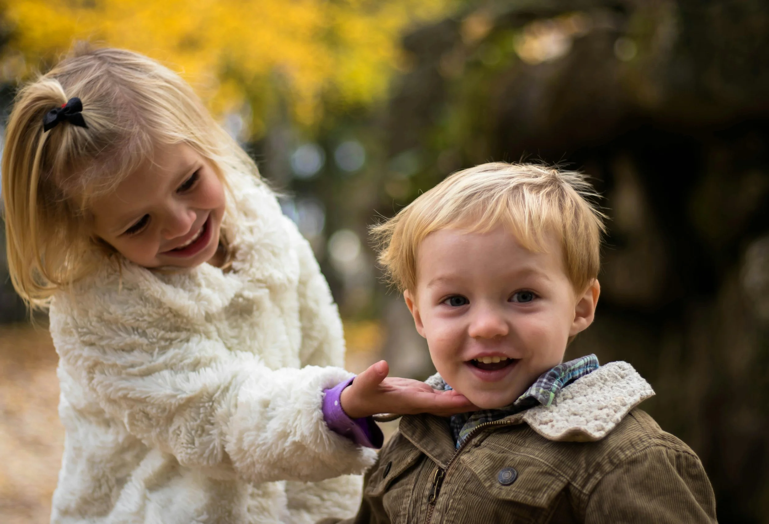 A young girl with blonde hair in a white fuzzy coat and black bow in her hair is playfully holding a boy's chin, who has blonde hair and is wearing a brown jacket with a fleece collar. They are outdoors with blurred autumn trees in the background.