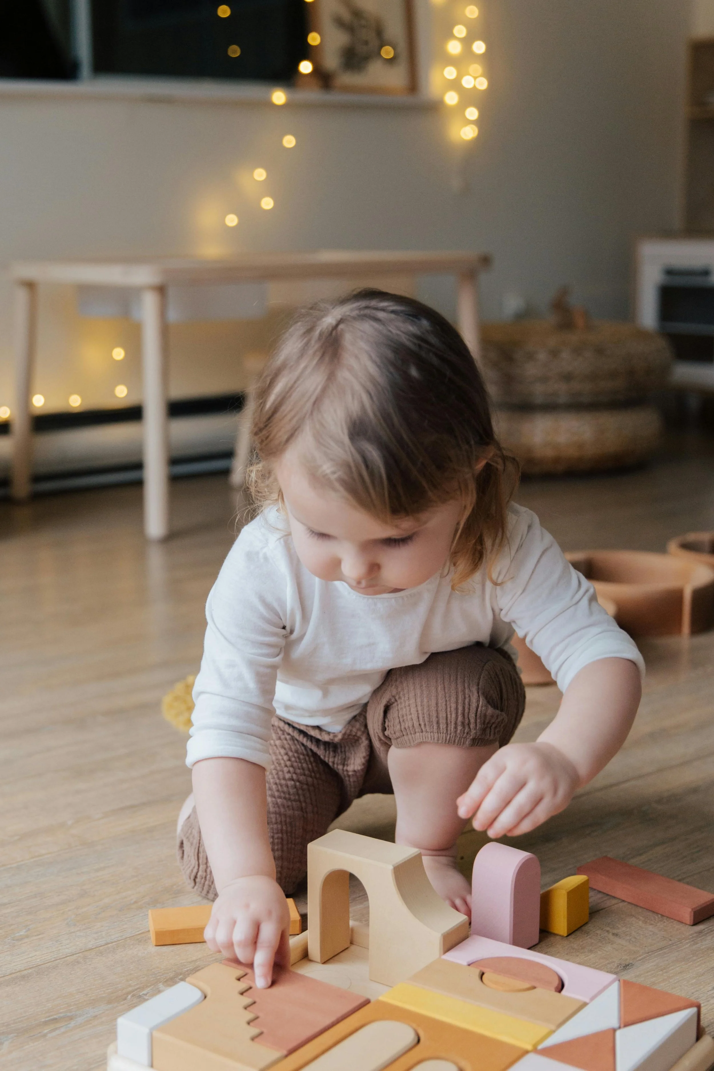 A young girl with brown hair playing with colorful wooden building blocks on the floor of a cozy room decorated with string lights.