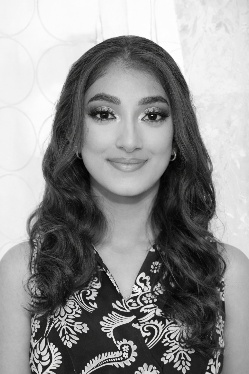 Black and white portrait of a young woman with wavy hair, wearing makeup and a patterned sleeveless top, smiling softly.