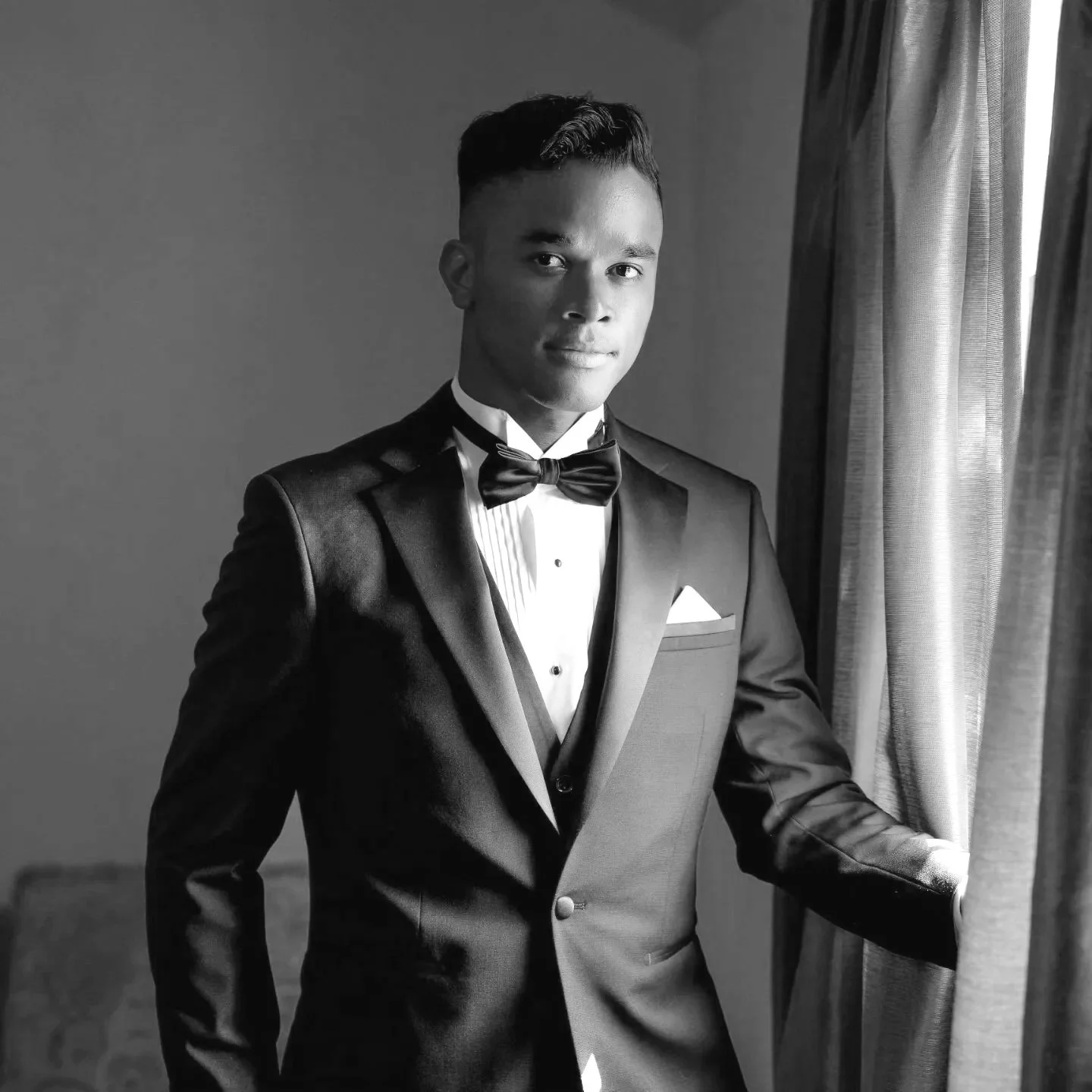 A young man in a tuxedo with a bow tie, standing near a window with curtains, looking at the camera, in a black and white photo.