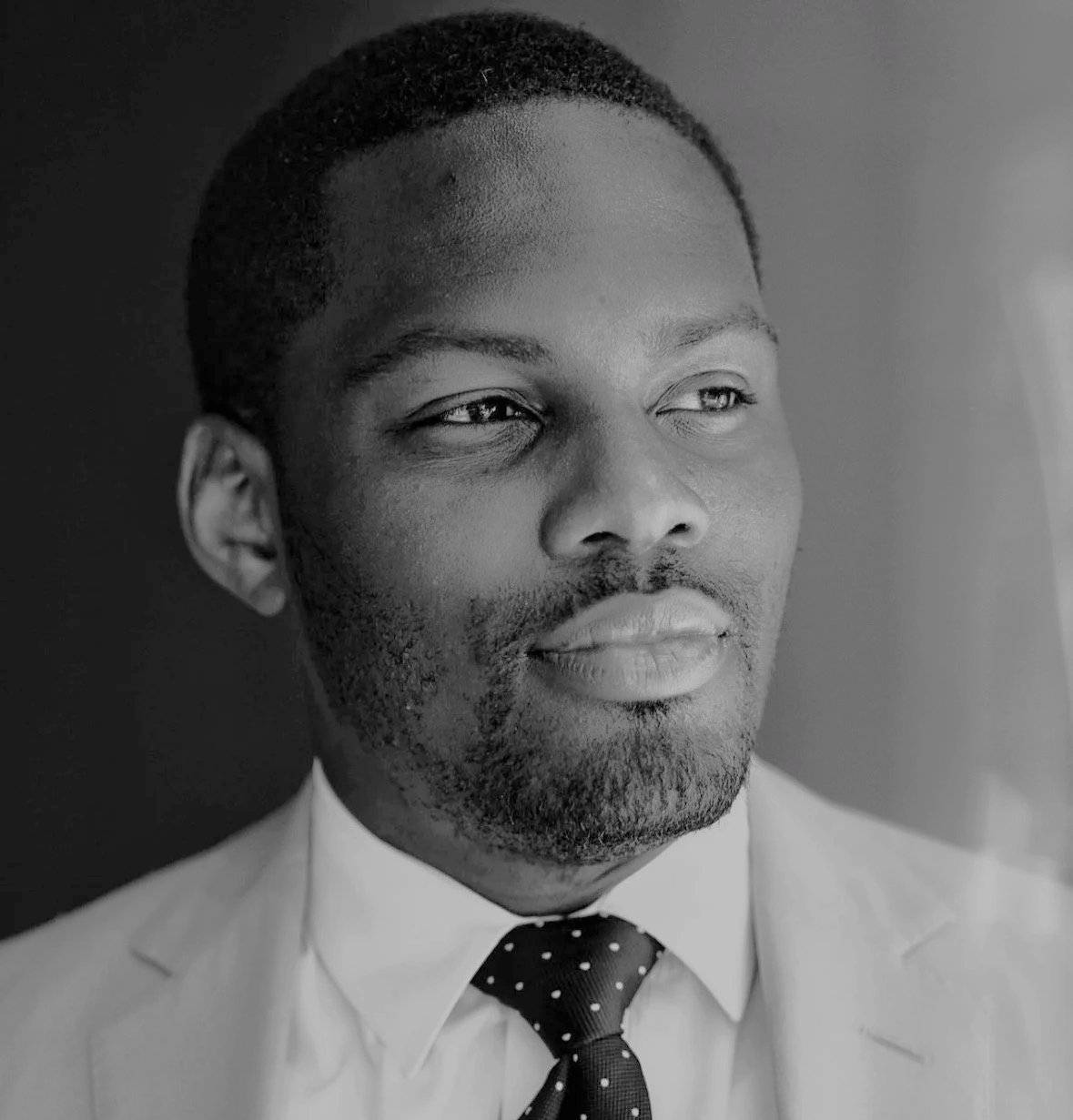 Close-up black and white portrait of a man wearing a suit and polka dot tie, looking thoughtfully to the side.