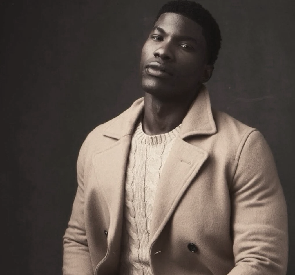 A young man with dark skin and short curly hair poses against a dark background, wearing a beige coat over a cable-knit sweater, with a slightly tilted head and a serious expression.