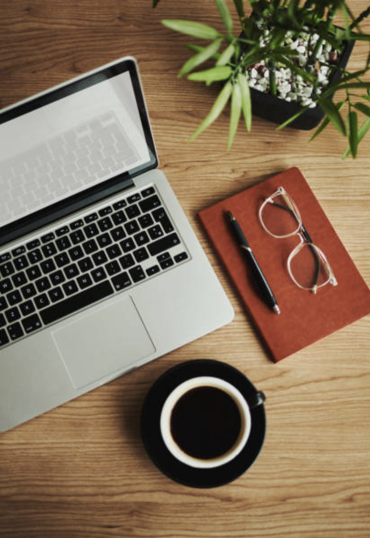 Desk with a laptop, eyeglasses, pen on a red notebook, a cup of coffee, and a potted plant on a wooden surface.