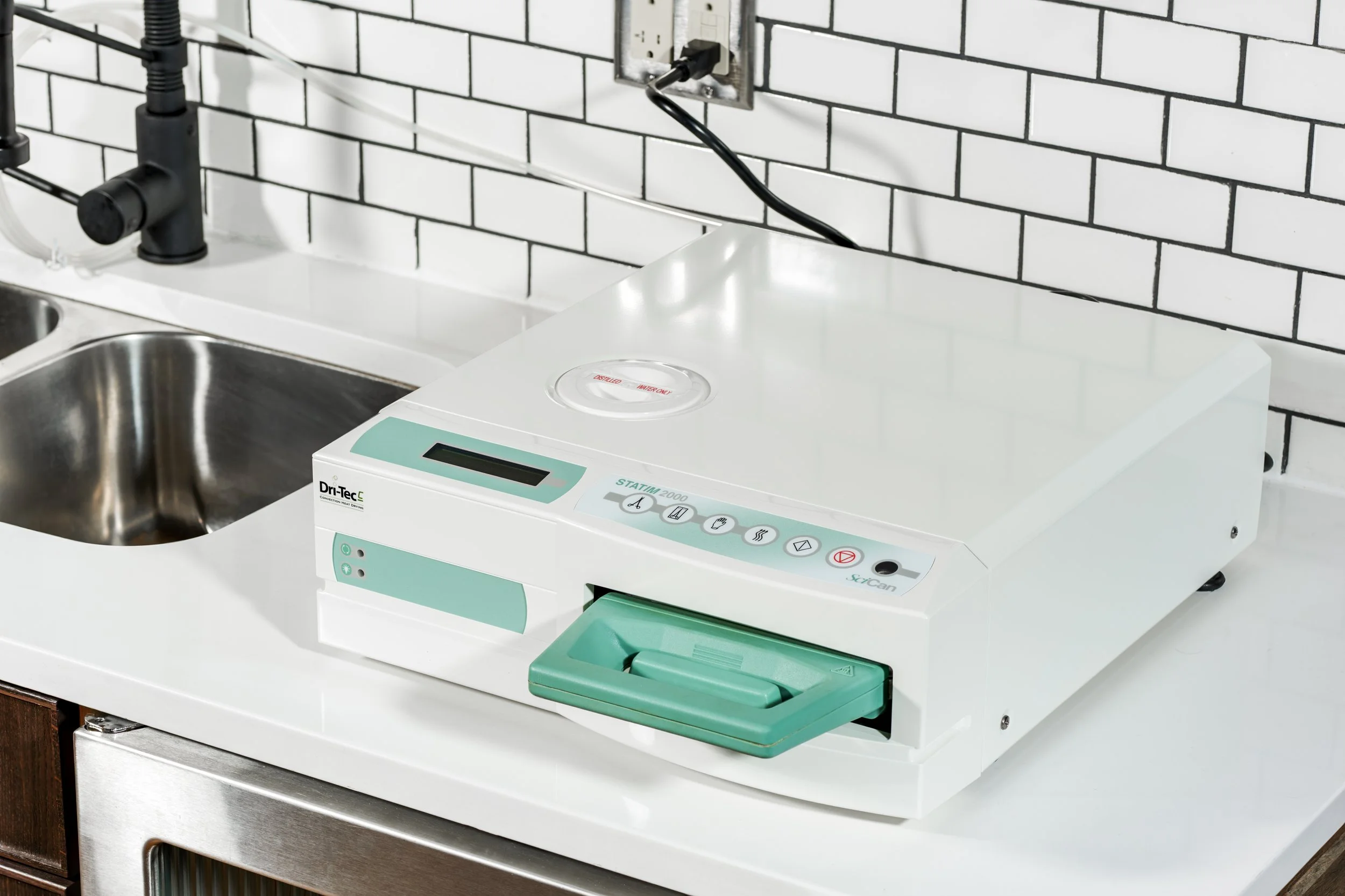 Medical sterilizer machine on a white countertop in a kitchen with a stainless steel sink and white brick backsplash.