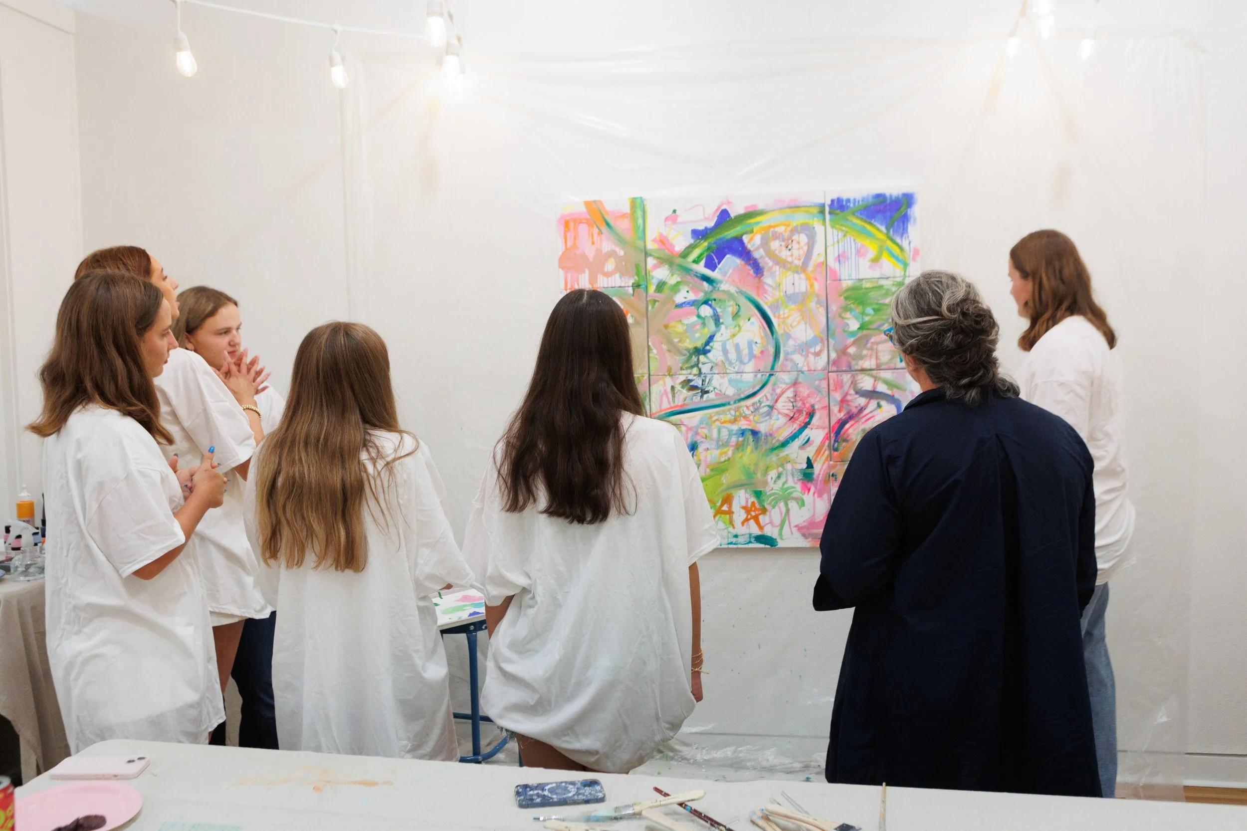 A group of women in white shirts and one woman in dark clothing standing in front of an abstract colorful painting on a white wall, observing and discussing the artwork in a gallery setting.