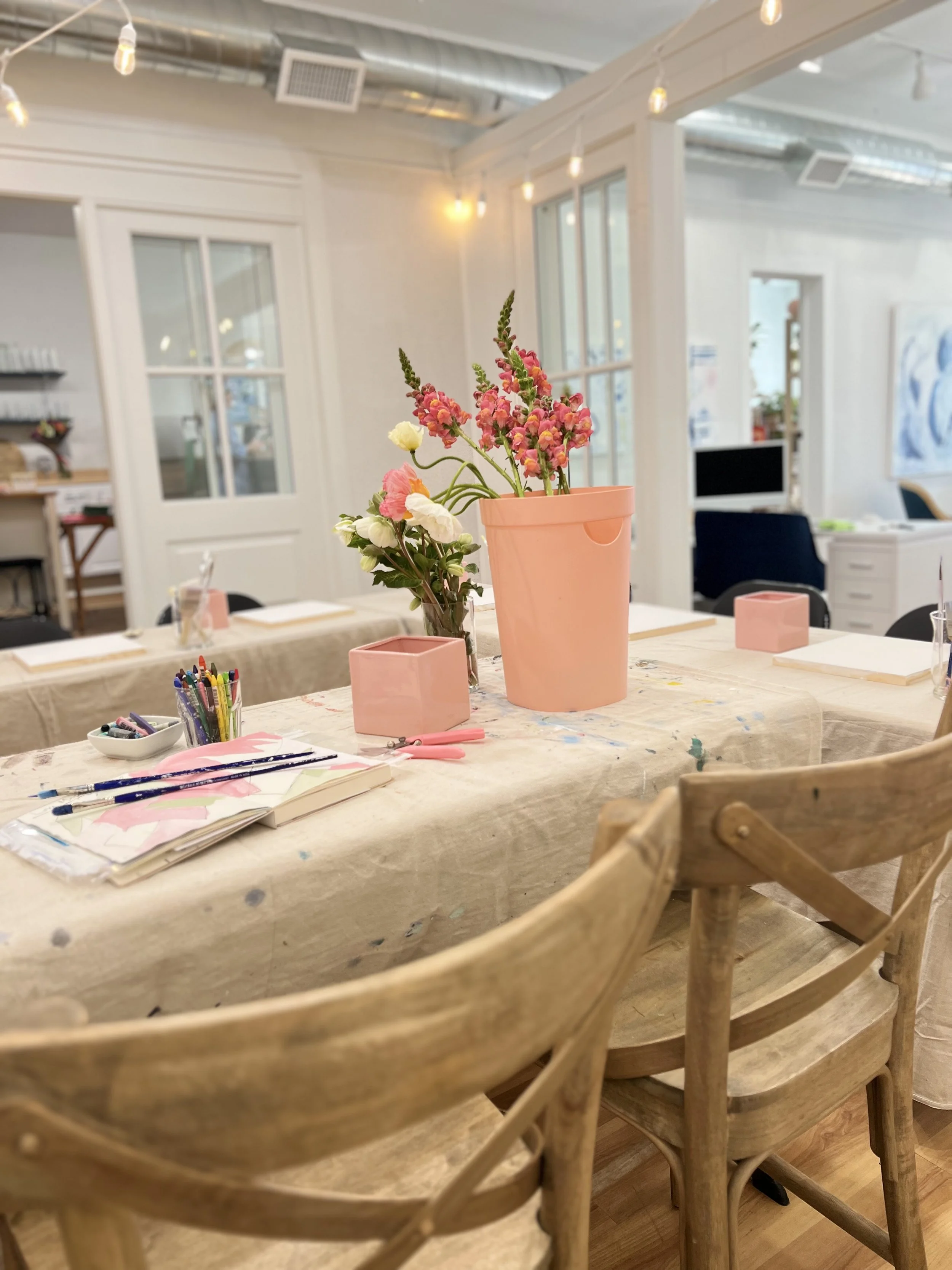 Art studio with wooden chairs, a table covered in a paint-stained cloth, art supplies, and pink flowers in vases.