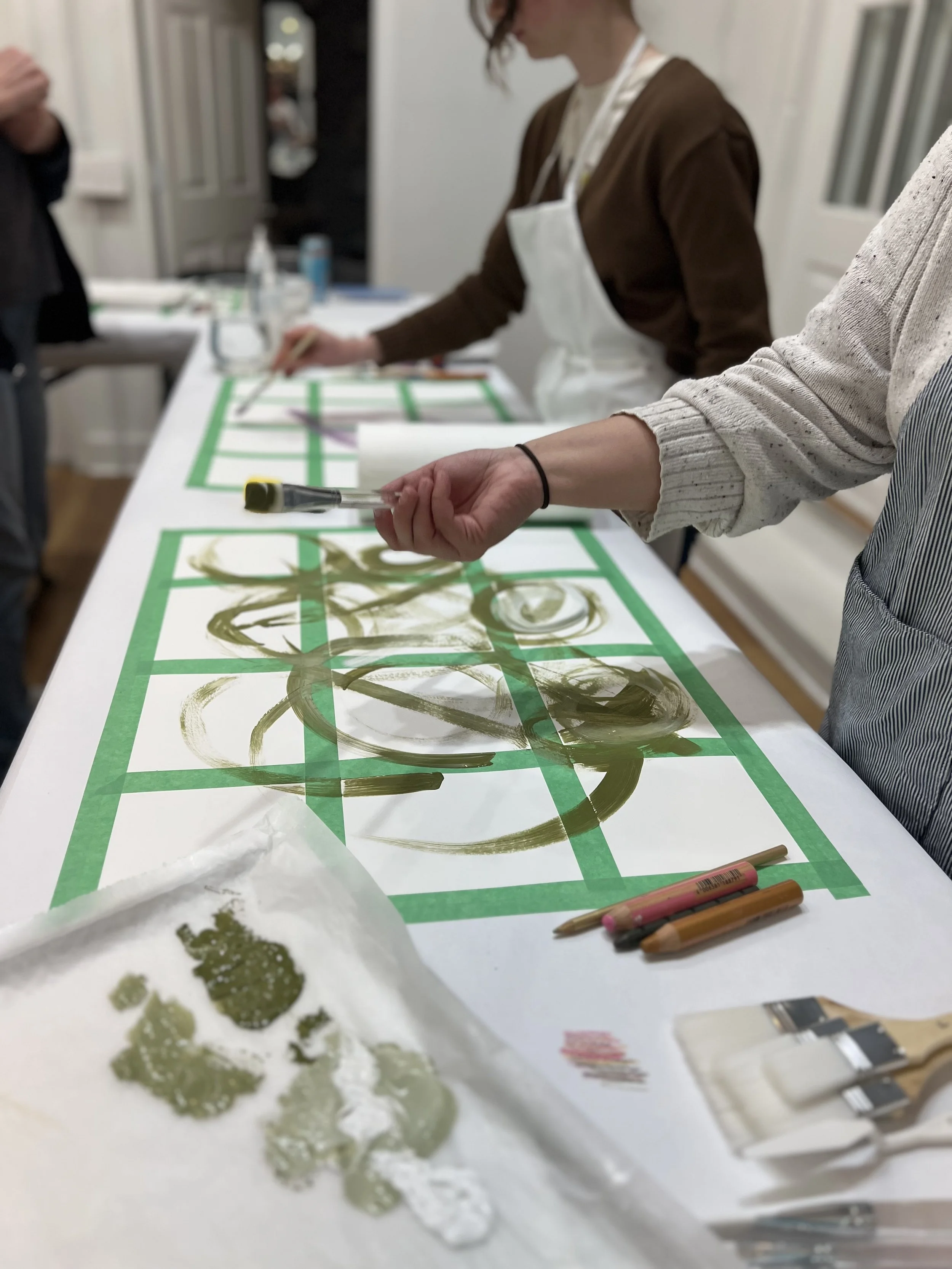 People painting abstract designs with green and white paint on paper, using brushes and pencils, in an art studio setting.