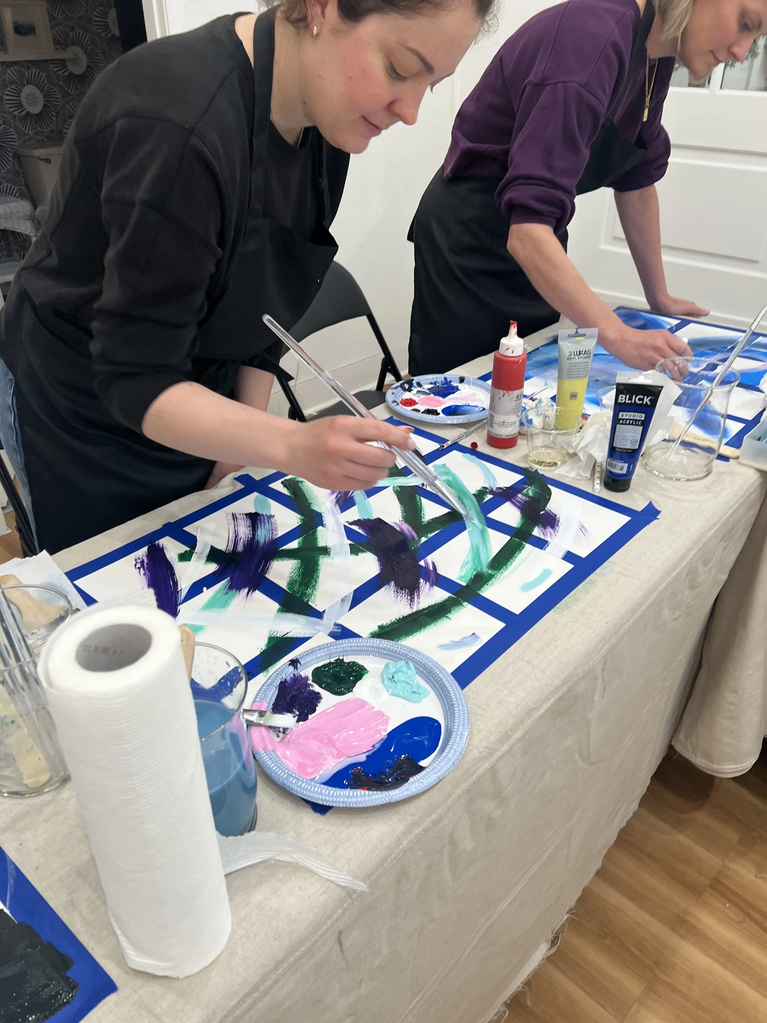 Two women are painting abstract designs on paper with colorful paints, standing at a table covered with art supplies including paint, brushes, and paper, in a room with a wooden floor.