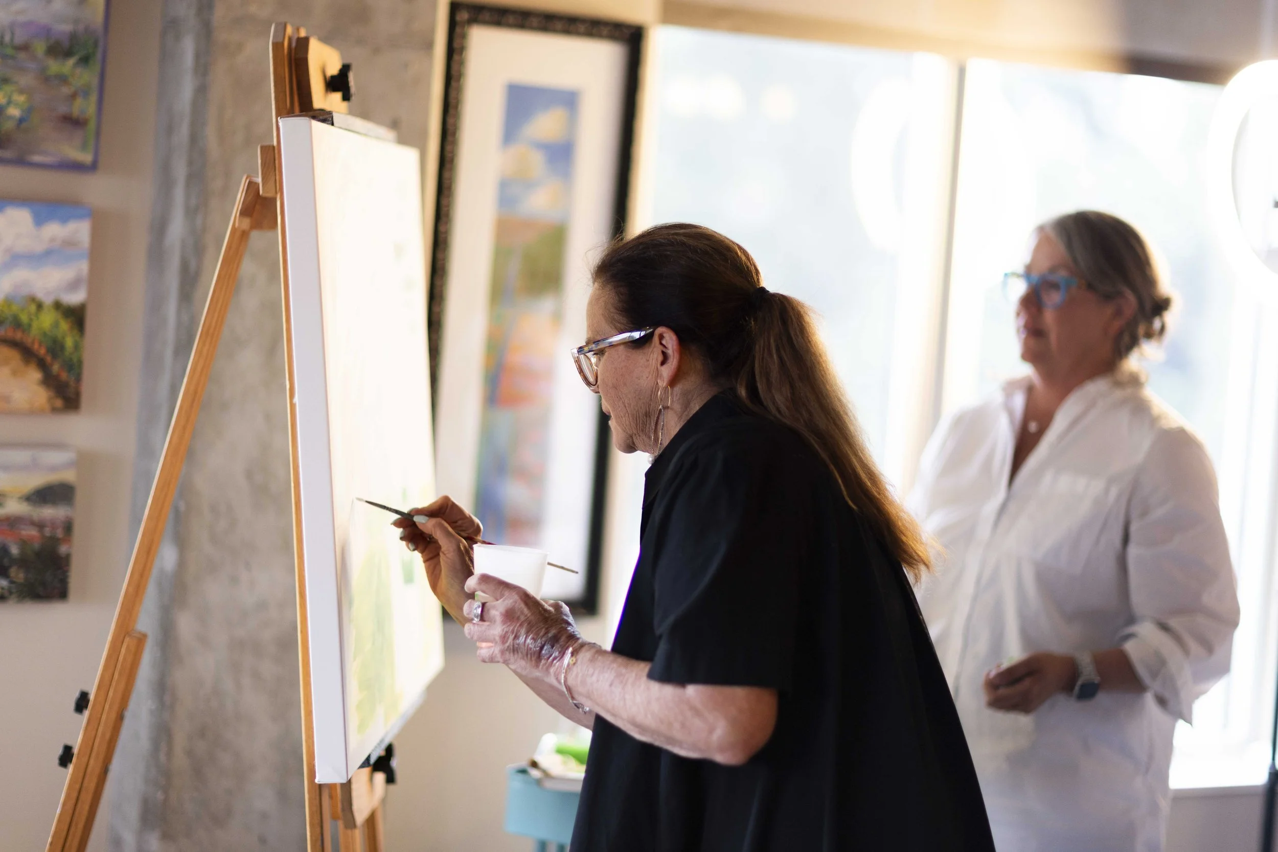 Two women in an art studio, one painting on a canvas and the other observing, with artwork hanging on the wall and sunlight coming through a window.