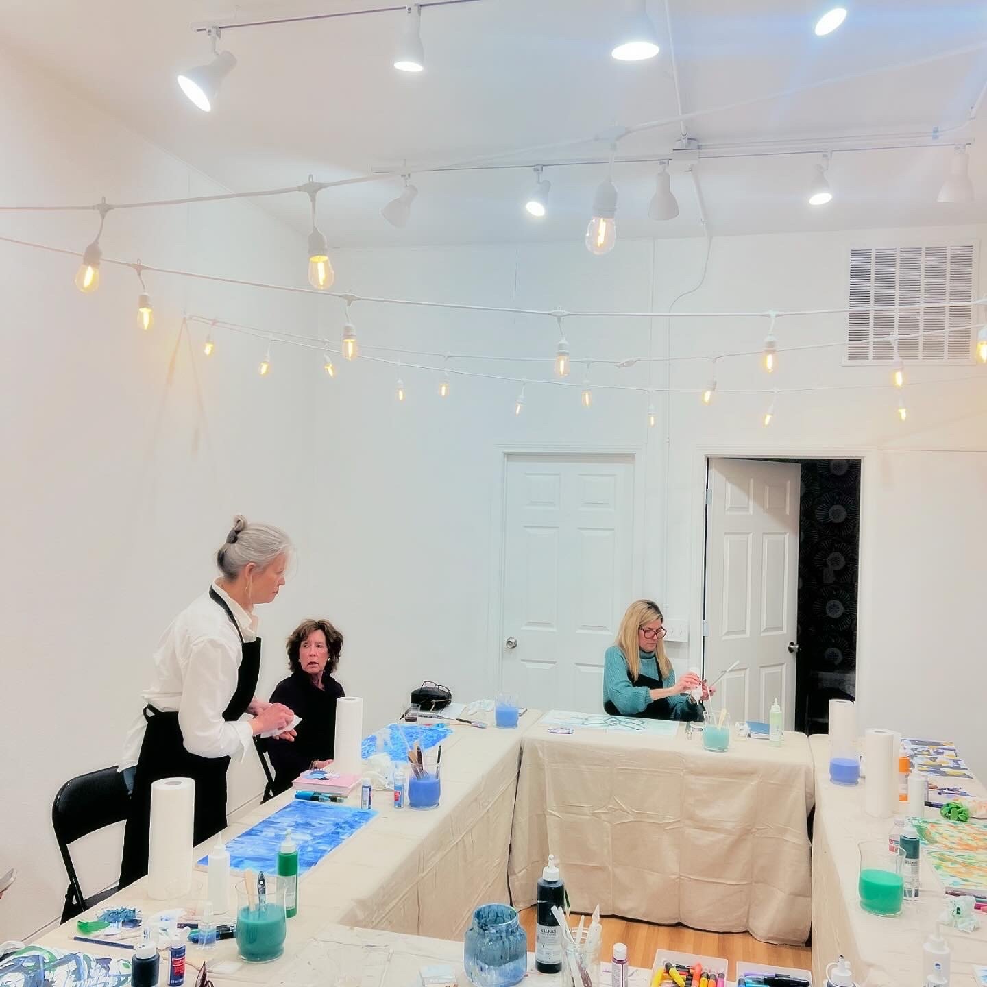 Art workshop with three women, tables covered in art supplies, and string lights overhead.