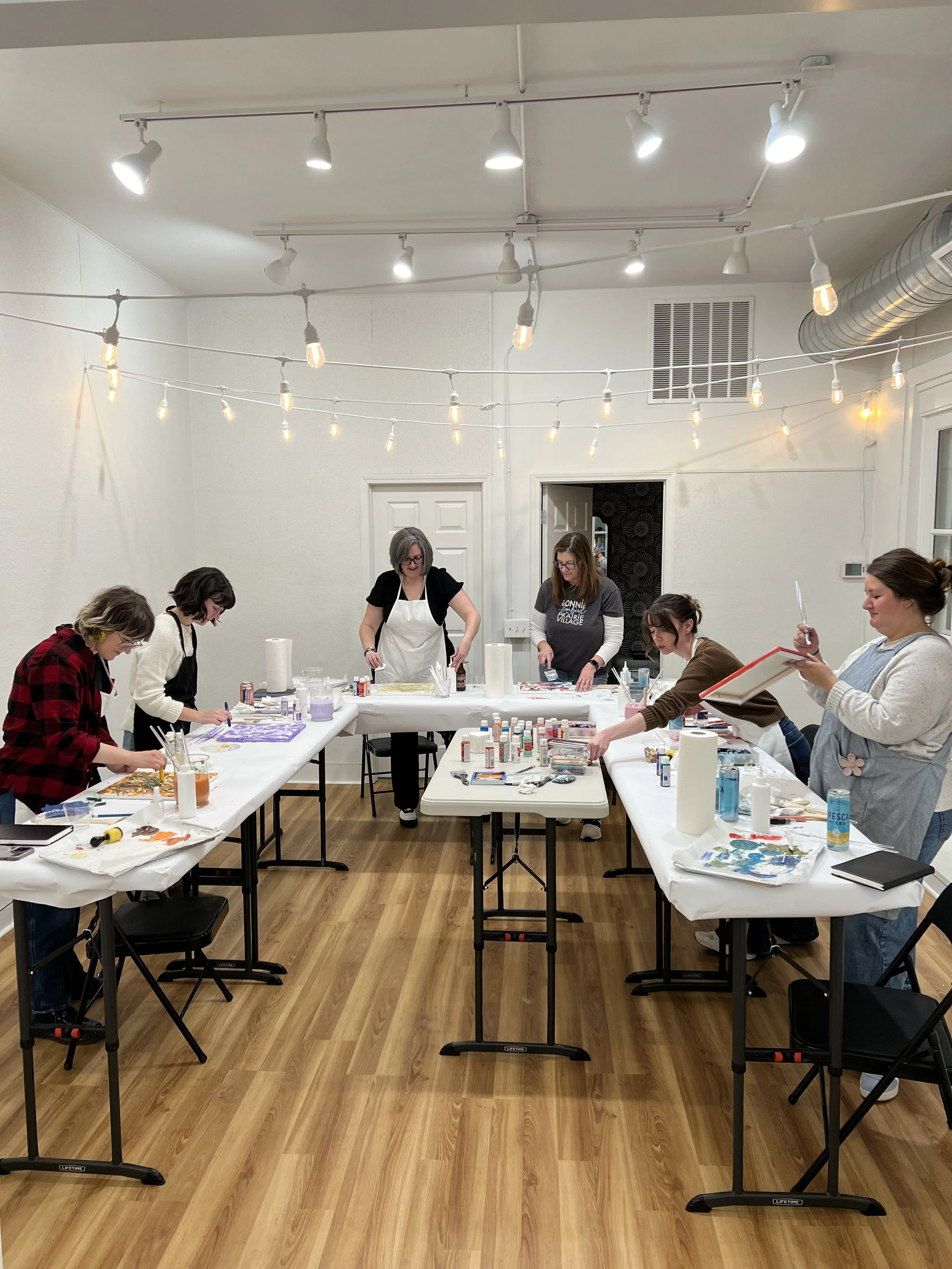 Group of women participating in an art class, working on creative projects at tables in a well-lit room.