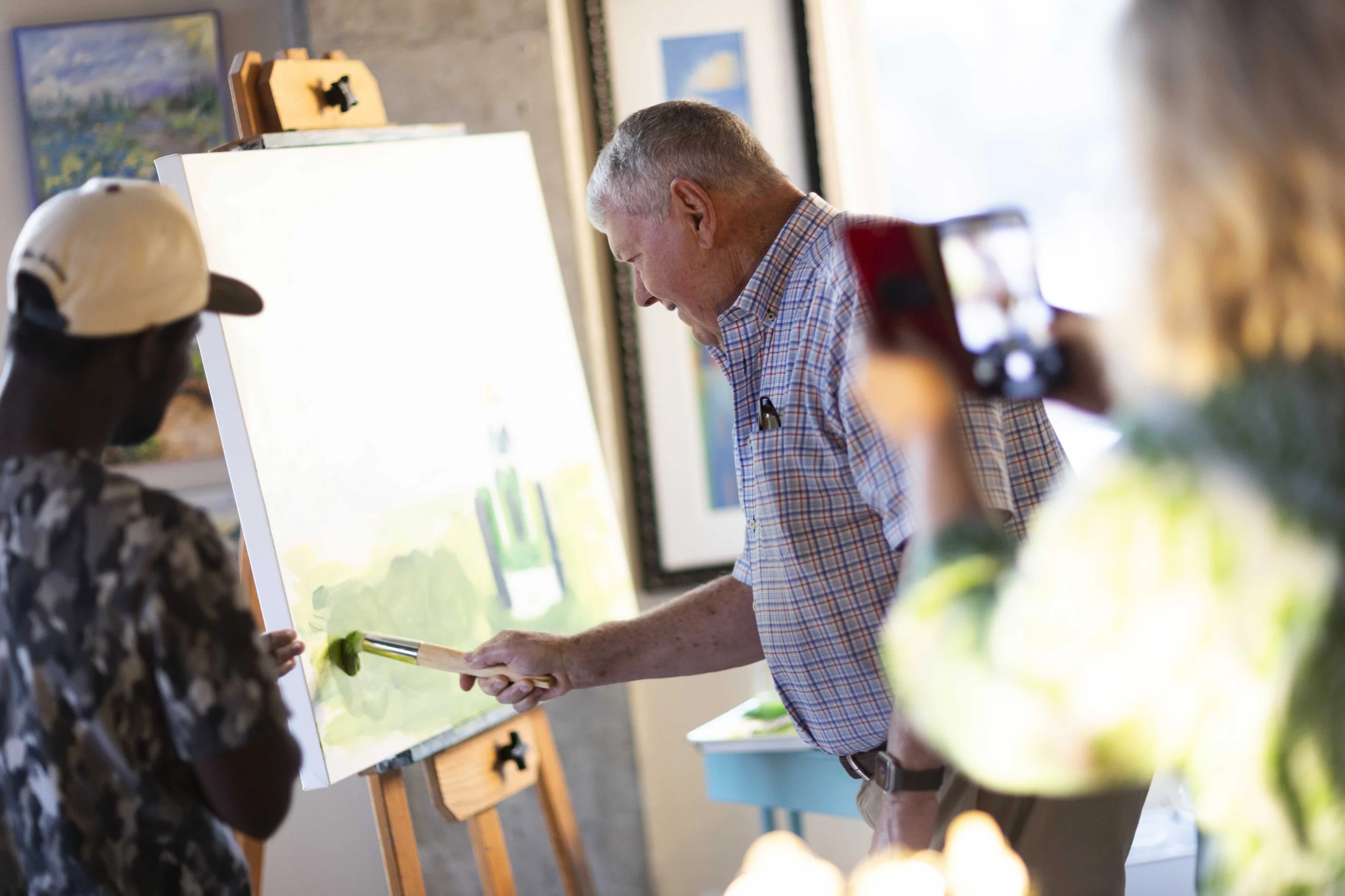 An elderly man with a short haircut wearing a checkered shirt uses a paintbrush to paint on a white canvas, while two observers watch, one person is taking a photo with a smartphone. The setting appears to be an art gallery or studio.